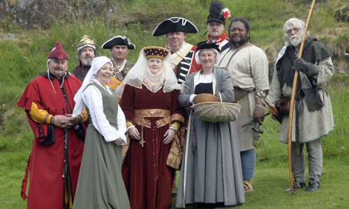 A group of historical reenactors in period costume stand in a group in front of a grassy hill.