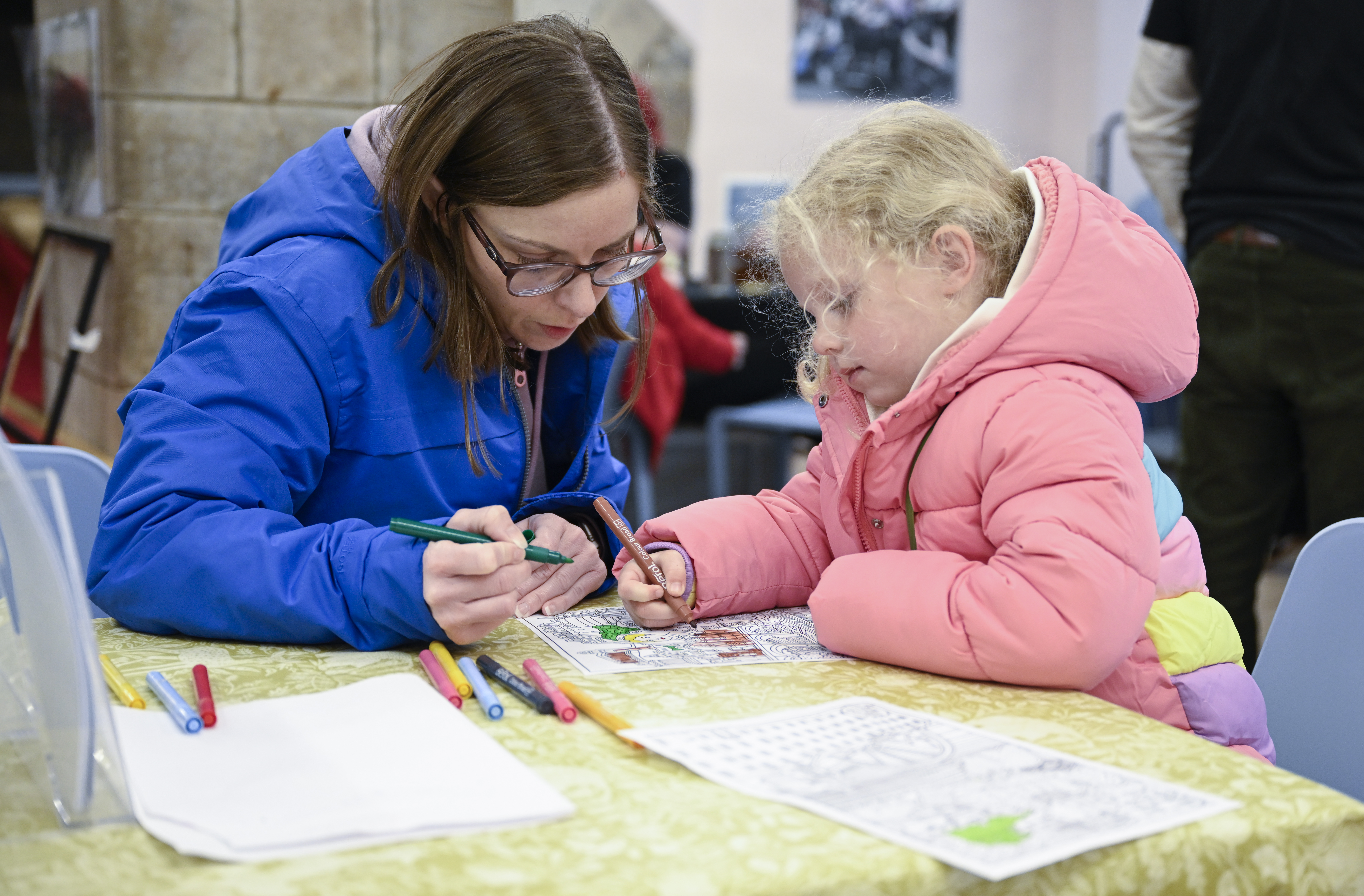 A woman and young girl sit at a table with scattered crayons, jointly filling in a colouring sheet.