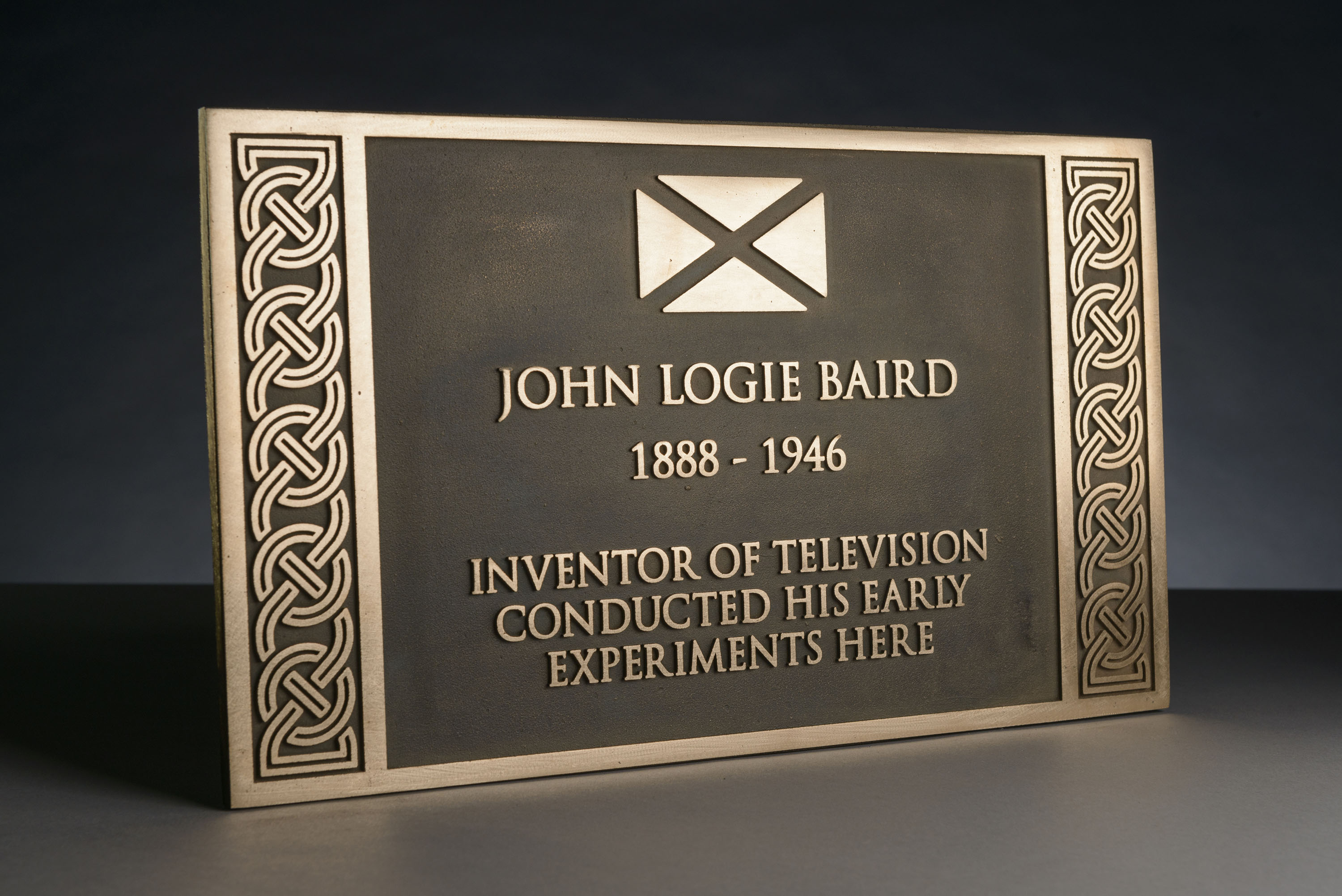 Studio photograph of a Historic Scotland commemorative plaque, shown at a slight angle against a dark gradient background. The rectangular plaque features a textured dark centre with a light border and vertical Celtic knot designs on both sides, and a raised Scottish saltire symbol at the top. The inscription reads: “John Logie Baird (1888–1946), inventor of television, conducted his early experiments here.”