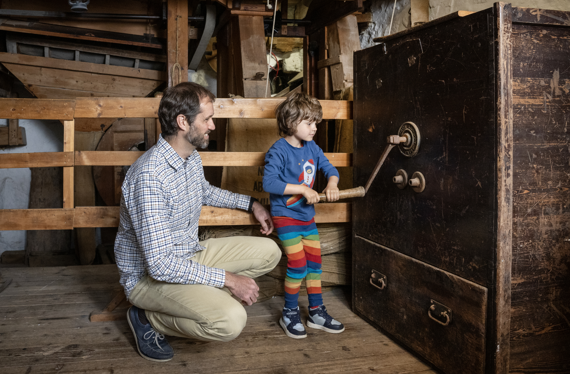 A child turning a large wooden handle on an old mechanical device inside the mill, with an adult kneeling nearby.