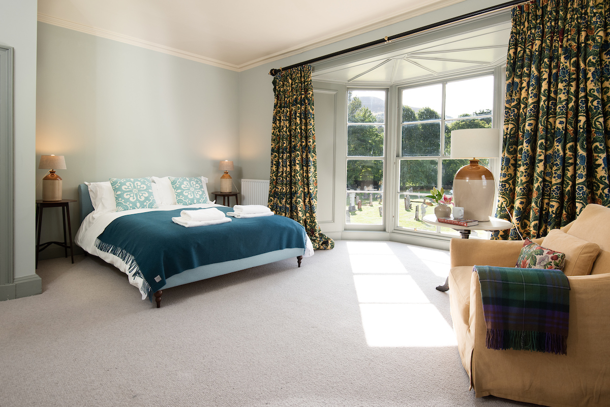 A large double bedroom, with light grey walls and carpet, a large, floor-to-ceiling bay window, and a large armchair 