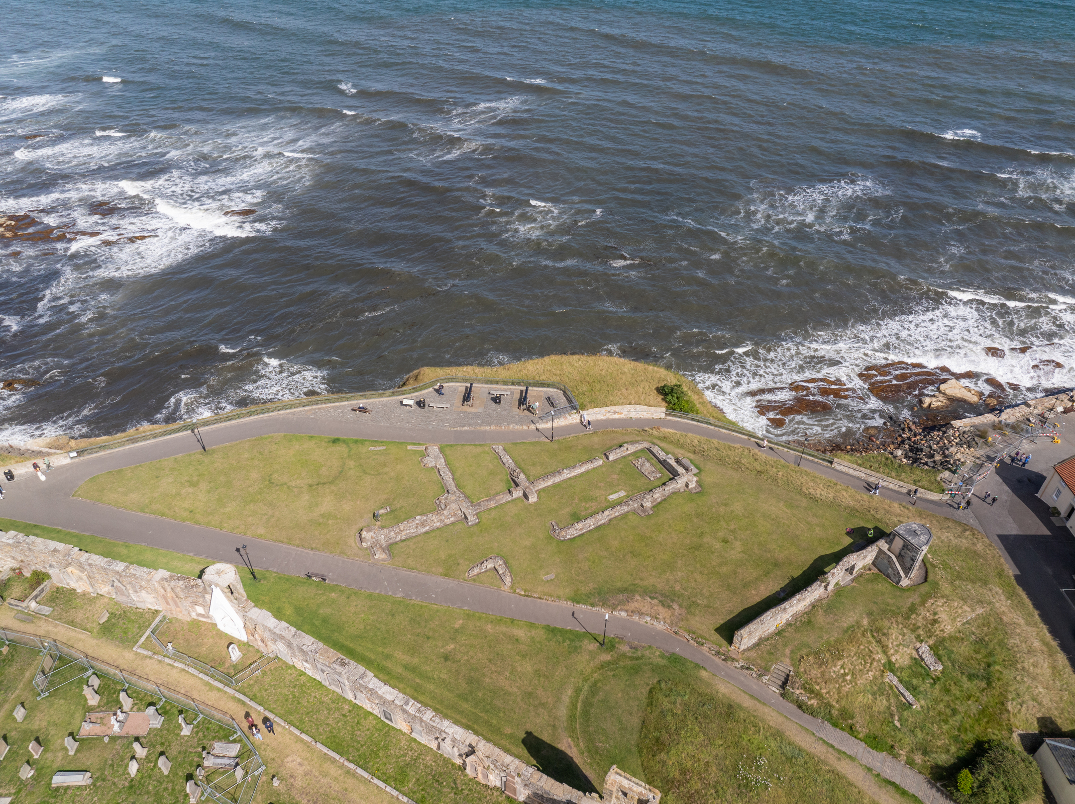 Overhead view of St Mary’s on the Rock ruins on a grassy clifftop, with the sea and rocky shoreline beside the site.