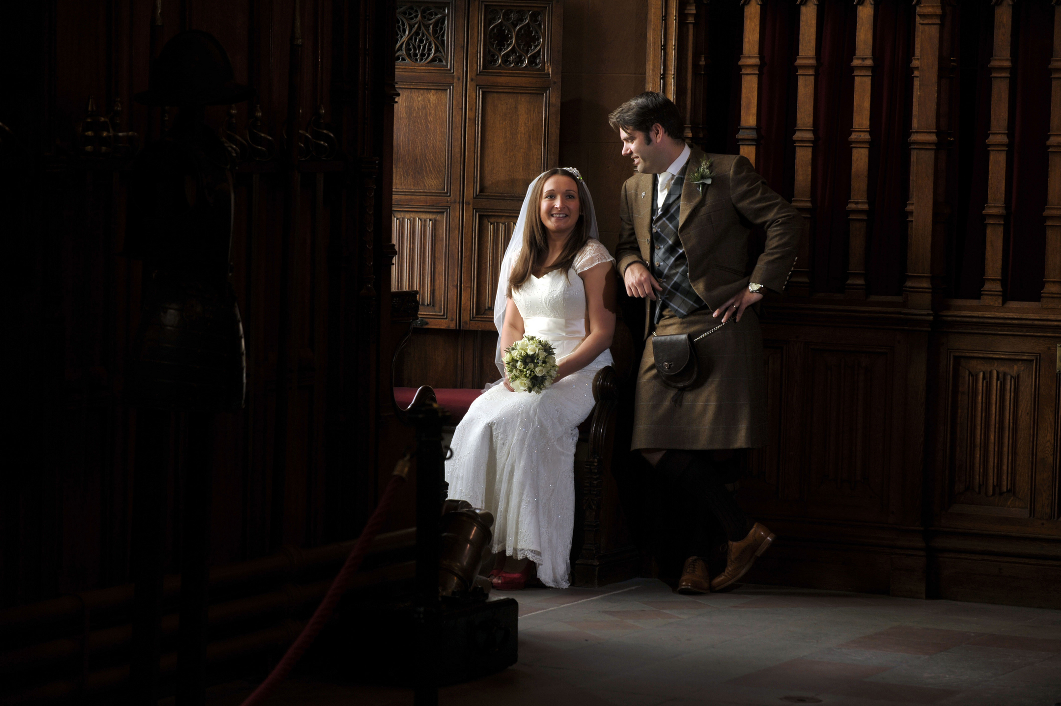 A bride sits next to her standing groom in a wood-paneled room. The two are lit by the light of a window out of shot. 