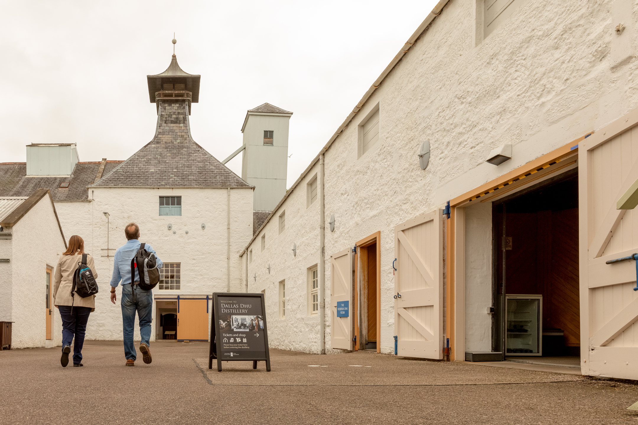 Visitors walk through the courtyard of Dallas Dhu distillery, surrounded by traditional whitewashed buildings and open warehouse doors leading into the historic site.