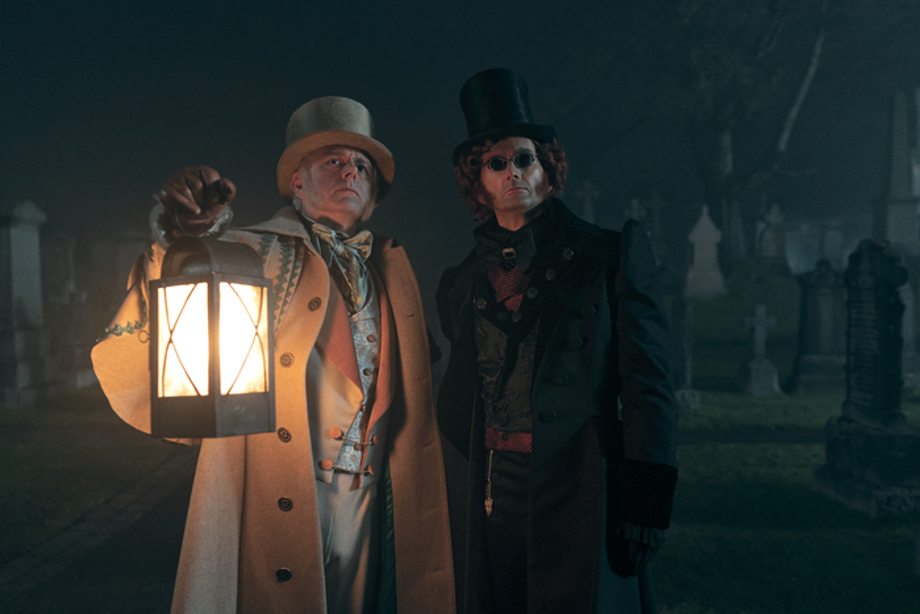 Two men in Victorian-style clothing stand in a foggy graveyard at night. One wears a pale coat and top hat and holds a glowing lantern; the other, dressed in dark clothing with a tall black hat and tinted glasses, stands beside him.