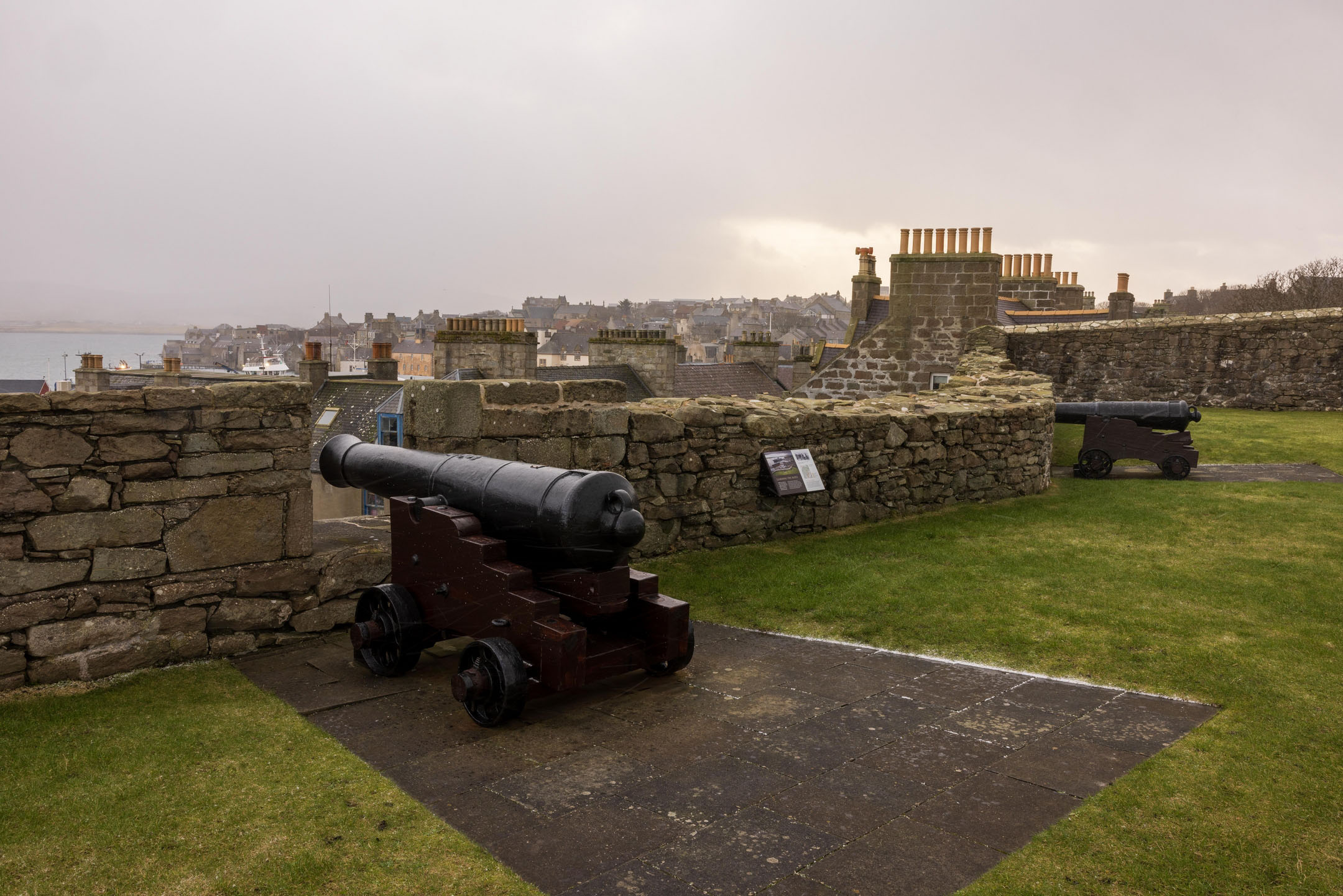 Cannons on the rampart overlooking town rooftops and harbour under overcast sky.