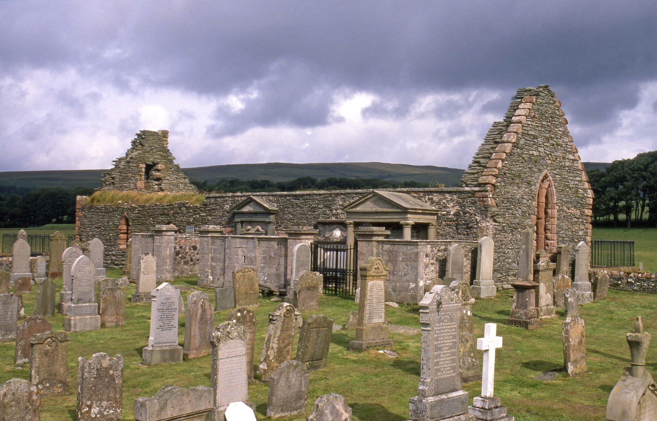 A ruined stone chapel surrounded by a proliferation of gravestones including medieval carved grave slabs and smaller white crosses.