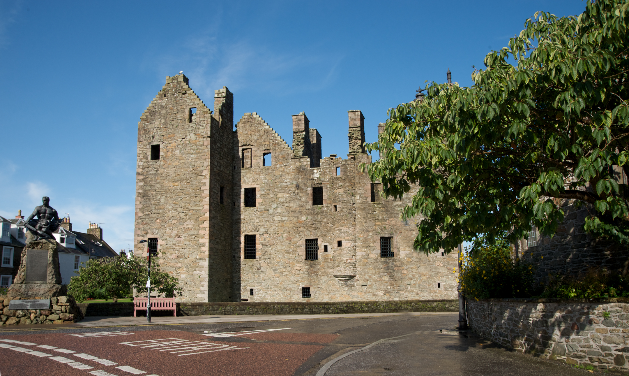 MacLellan’s Castle viewed from across a road, with a statue on a plinth to the left and a leafy tree partially framing the right side.