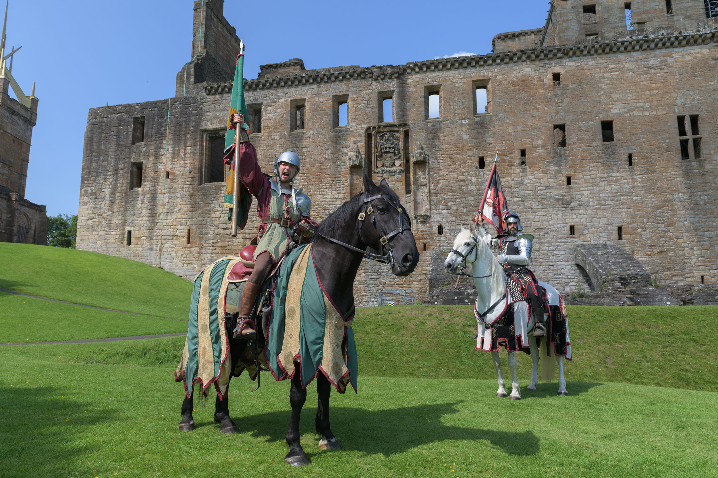 Two men in medieval knight costumes on horseback pose holding flags above their heads in front of a large stone castle.