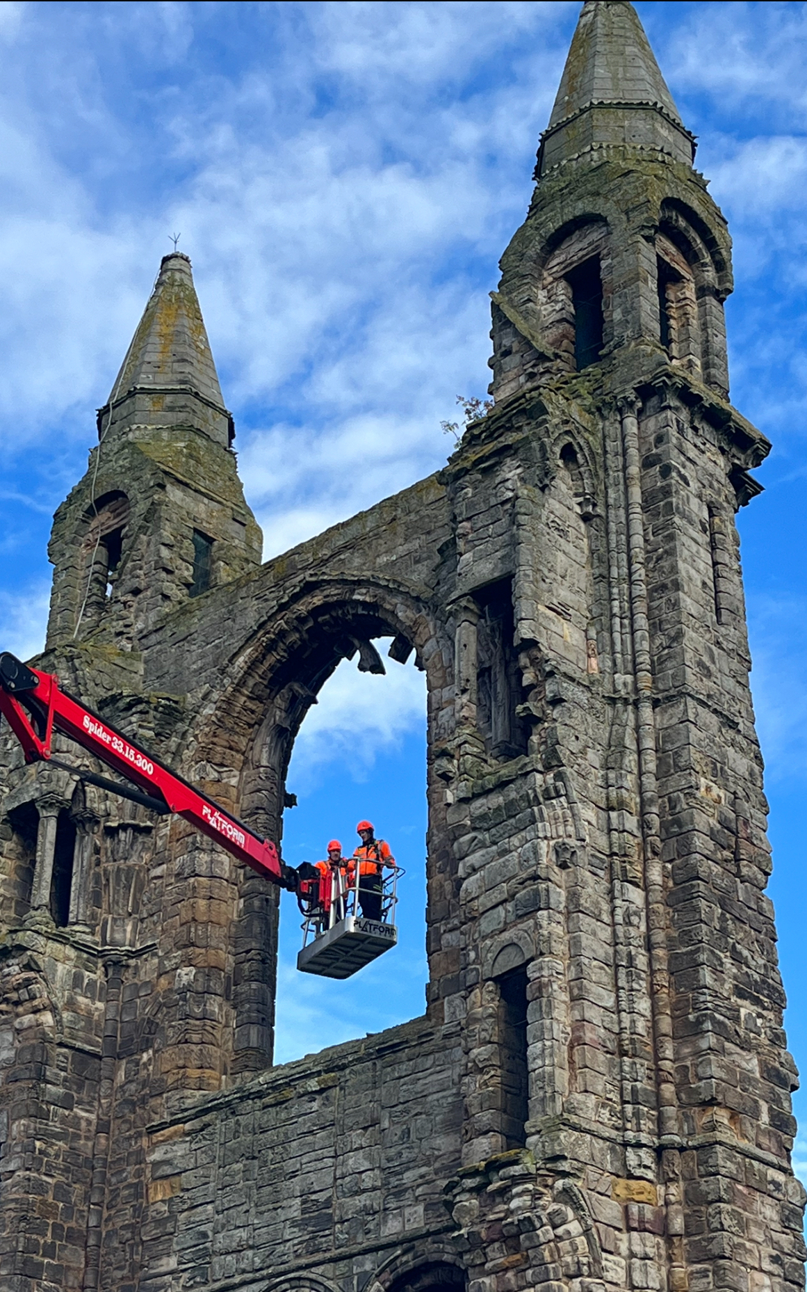 Stone ruins of St Andrews Cathedral with two tall towers, viewed against a blue sky, with a red cherry picker raised inside an arched opening.