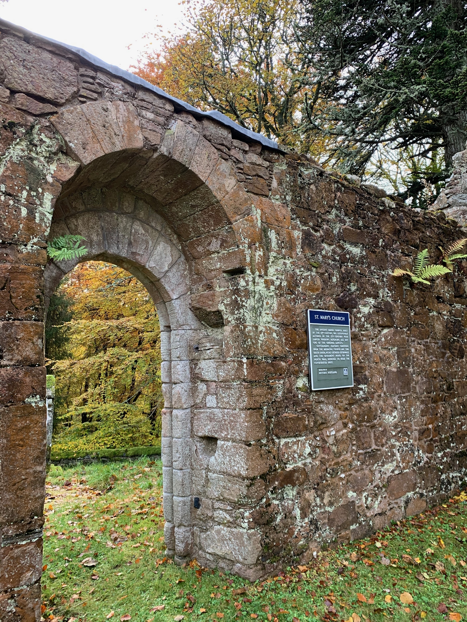 stone wall with an arched doorway and an iron plaque reading "St Mary's church. The ancient parish church, built in the 13th century, was extensively altered in the 17th century with larger windows replacing all but one of the original lancets. Outstanding features surviving from the mediaeval church are this south door, richly detailed externally, and the sacrament house on the north wall, designed to hold the consecrated host."