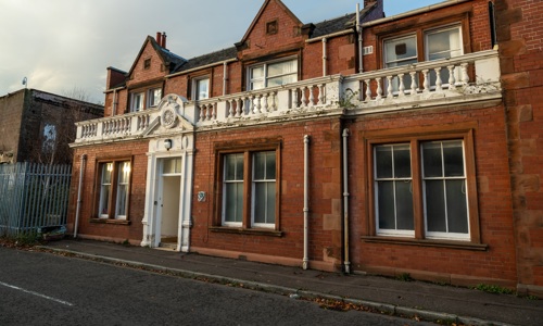 Historic two storey red brick building. A painted doorway provides main entrance, and decorative white balustrade runs along the top of first storey