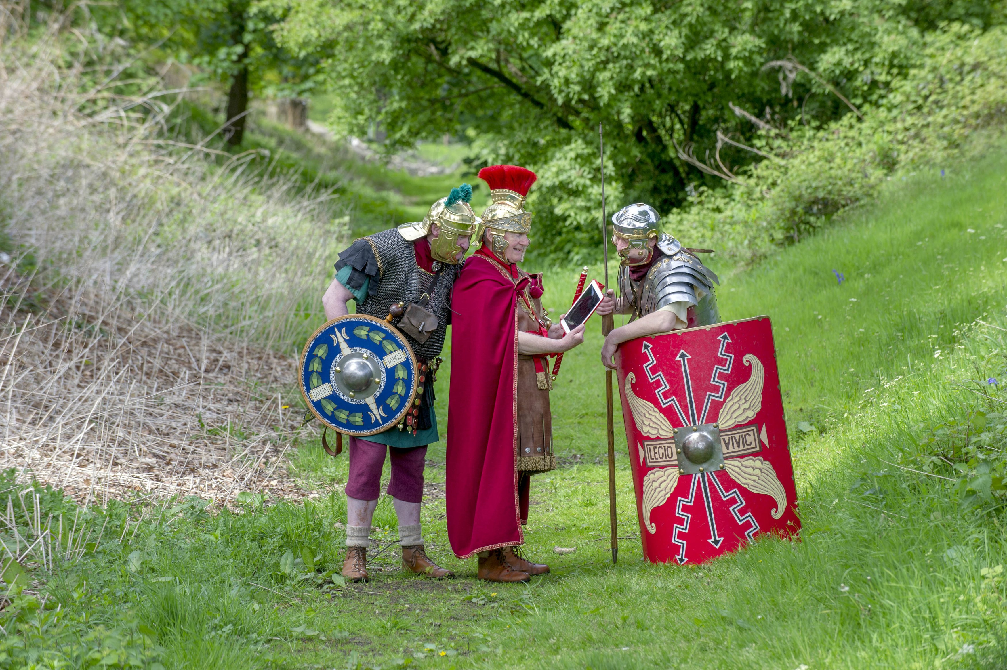 Three costumed re-enactors dressed as Romans with red capes, armour and a rectangular shield. They standing in a grassy knoll, the remains of an ancient Roman fortification. As an amusing paradox, one of the Romans is holding a modern tablet device, and showing his companions the contents on screen. 
