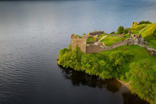 A ruined tower of a castle on a rocky promontory surrounded on two sides by water. Behind the tower is the remains of the other castle buildings and its walls.