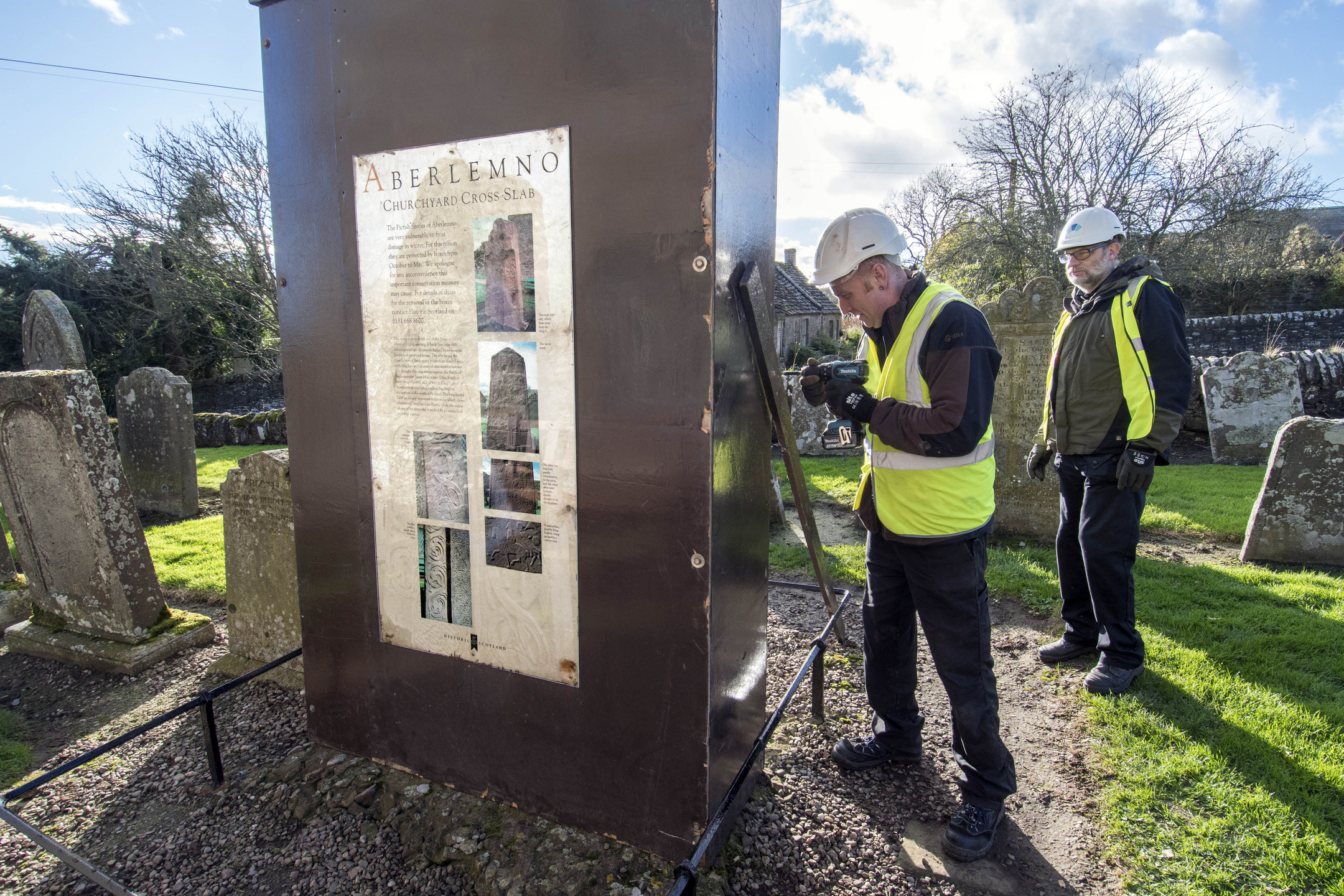 Workers in a churchyard place a wooden box around a carved stone to protect it during the winter. A panel with pictures and information about the stone is on the side of the box.