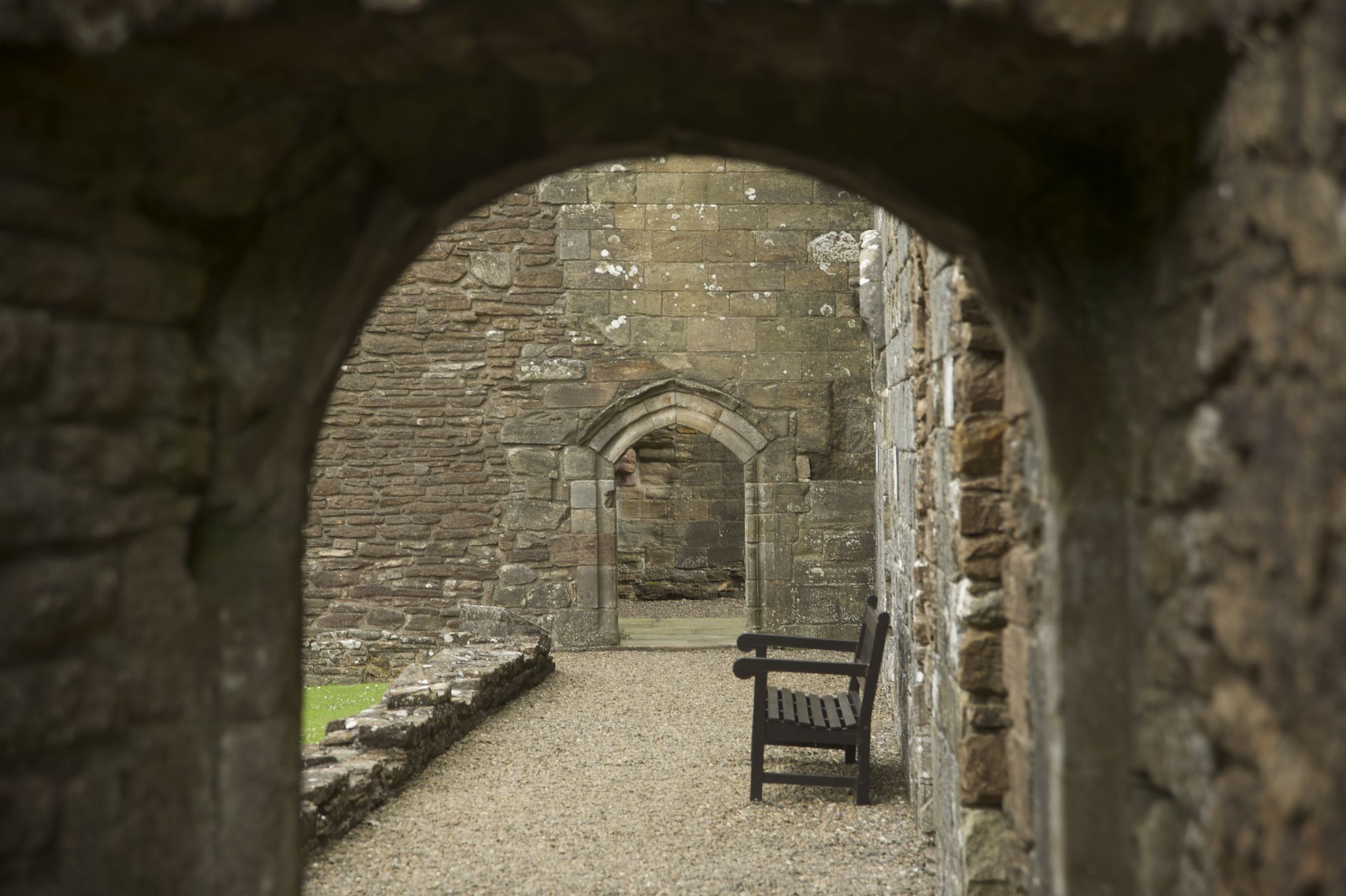 A view through a stone archway inside Crossraguel Abbey, showing a wooden bench along a gravel path and another small arch ahead.