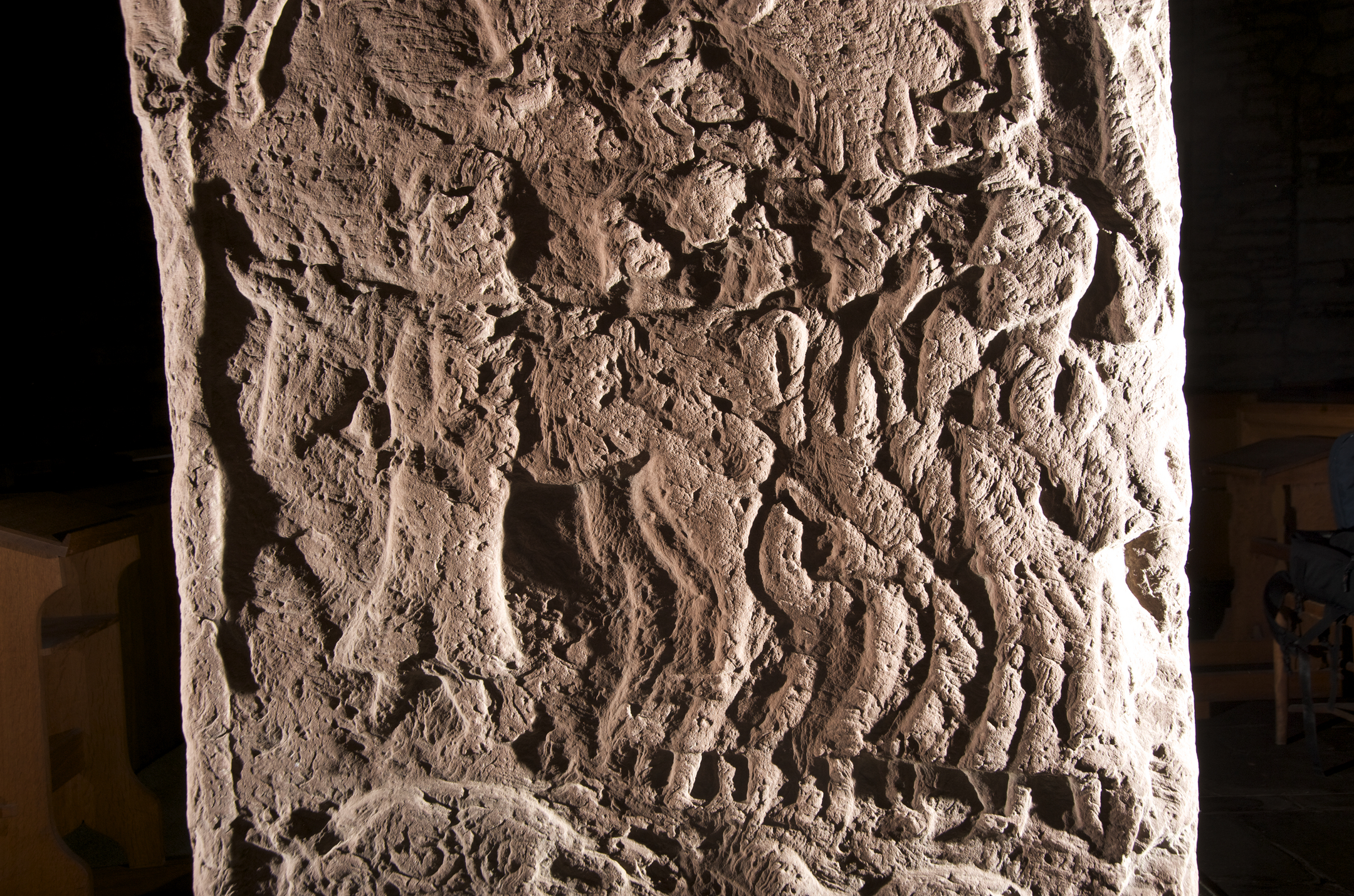 Close-up of an ancient sculptured stone showing detailed carvings of human figures and horses in relief. The surface is weathered, with intricate patterns and outlines depicting a scene of movement, possibly a procession or battle. The stone has a rough texture and is illuminated from the side, highlighting the depth of the carvings.