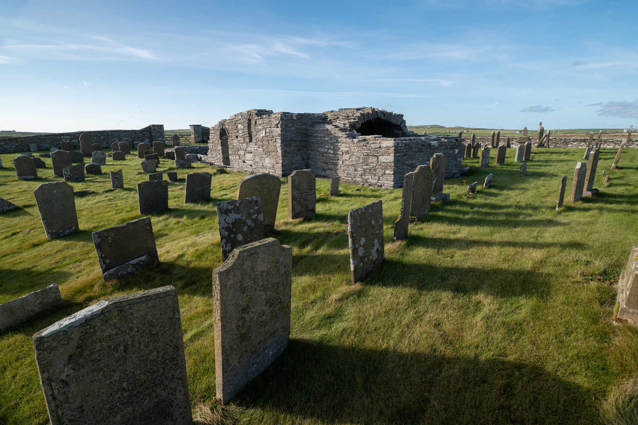 View across the graveyard toward the stone remains of Westside Church, with rows of gravestones casting long shadows on the grass.