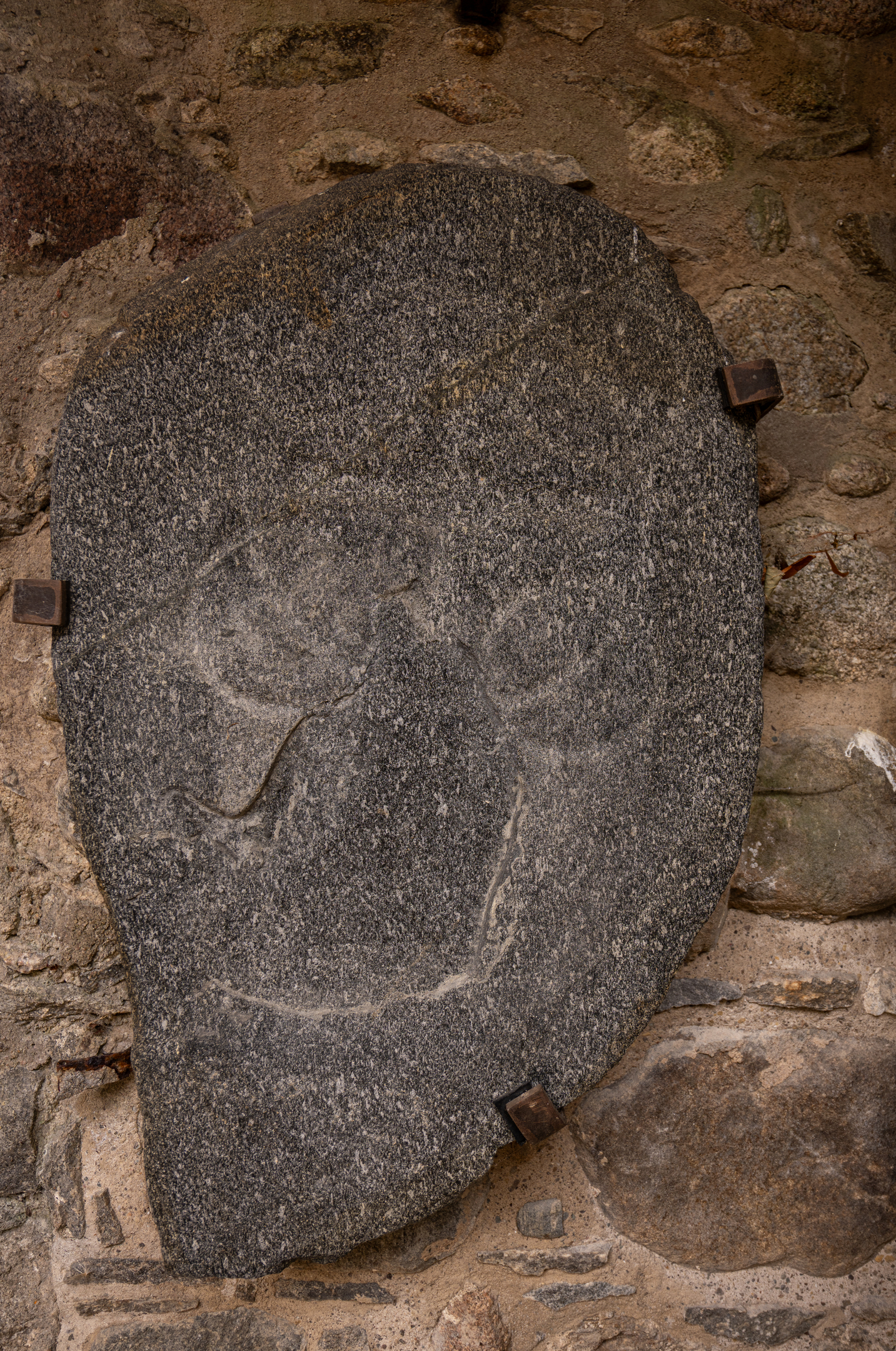 Oval-shaped Pictish stone mounted on a wall with metal brackets, displaying a simple incised cross with equal arms and angular terminals. The carving is shallow and weathered, contrasting with the rough stone surface.