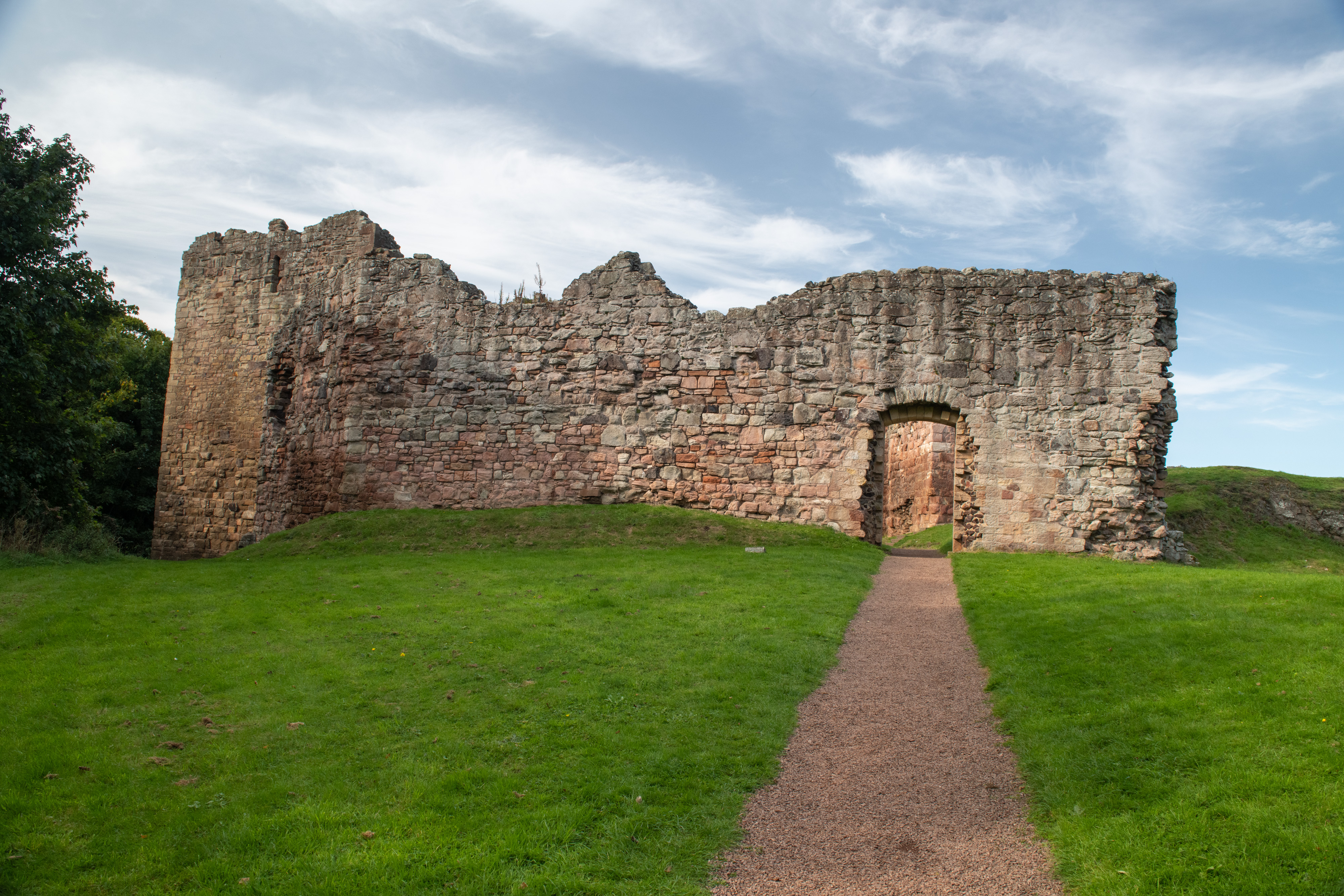 A weathered stone wall of Hailes Castle with a central arched doorway, standing on a grassy mound under a bright blue sky with wispy clouds. A gravel path leads directly to the entrance.