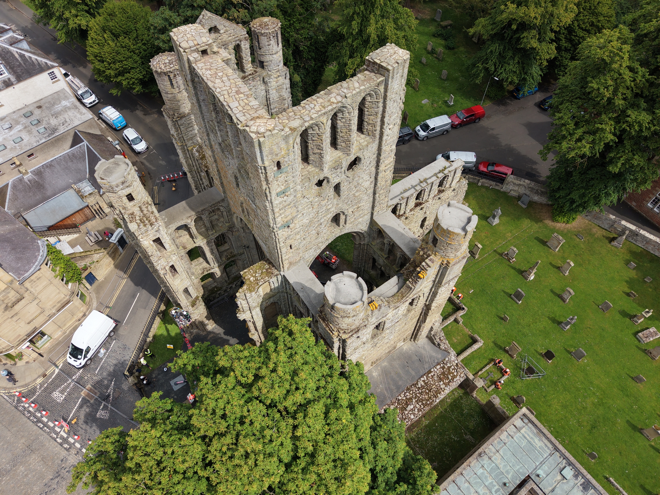 Aerial view of a large stone church ruin with a tall square tower and partial surrounding walls, seen from above through tree cover. The historic building sits beside a road with several parked vehicles, while a green churchyard with scattered gravestones extends to the right. 