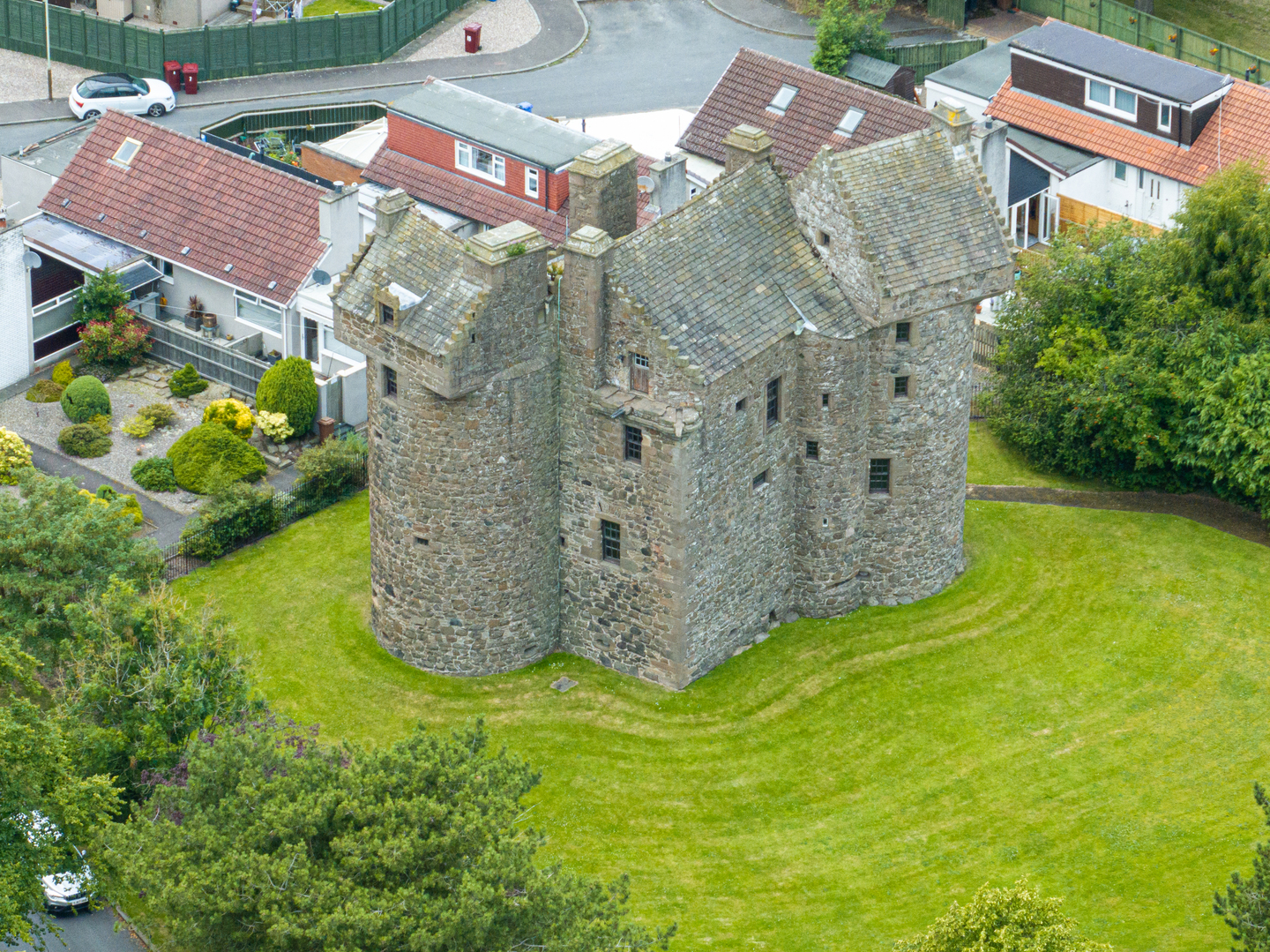 A stone castle, with an unusual two round towers as part of it. There is grass and trees surrounding it, with a road in the background. 