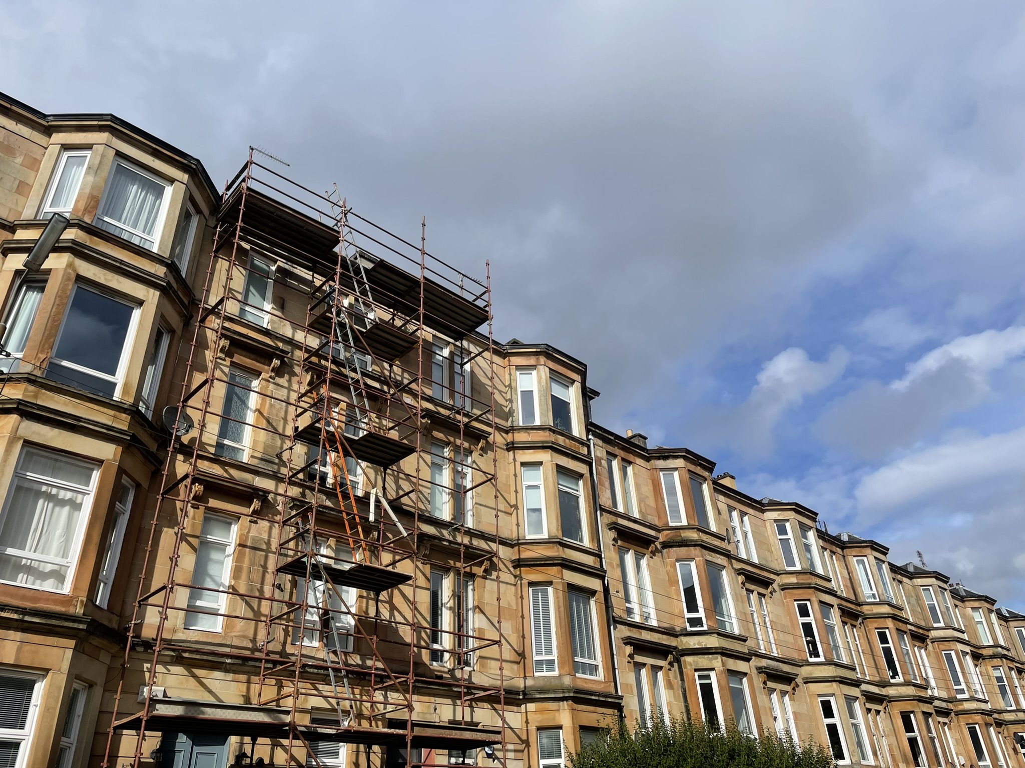 Traditional sandstone tenement building with multiple bay windows and a scaffold tower erected along the front façade for maintenance or repair work. The scaffolding extends from ground level to the roofline and includes ladders and working platforms. The terrace continues along the street, with neighbouring buildings of similar architectural.