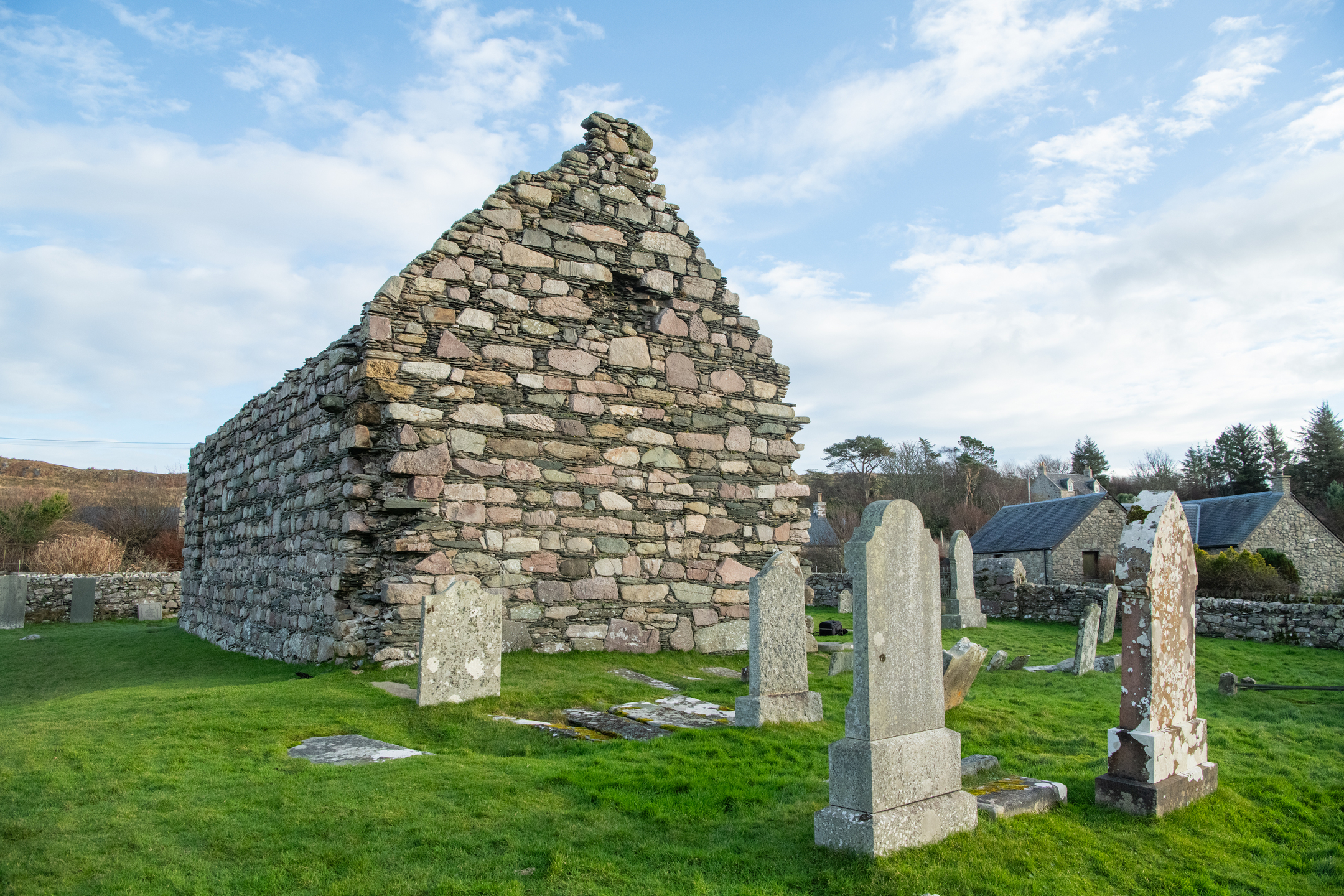 A gable end of a ruined stone chapel rising above a grassy graveyard with several weathered headstones.