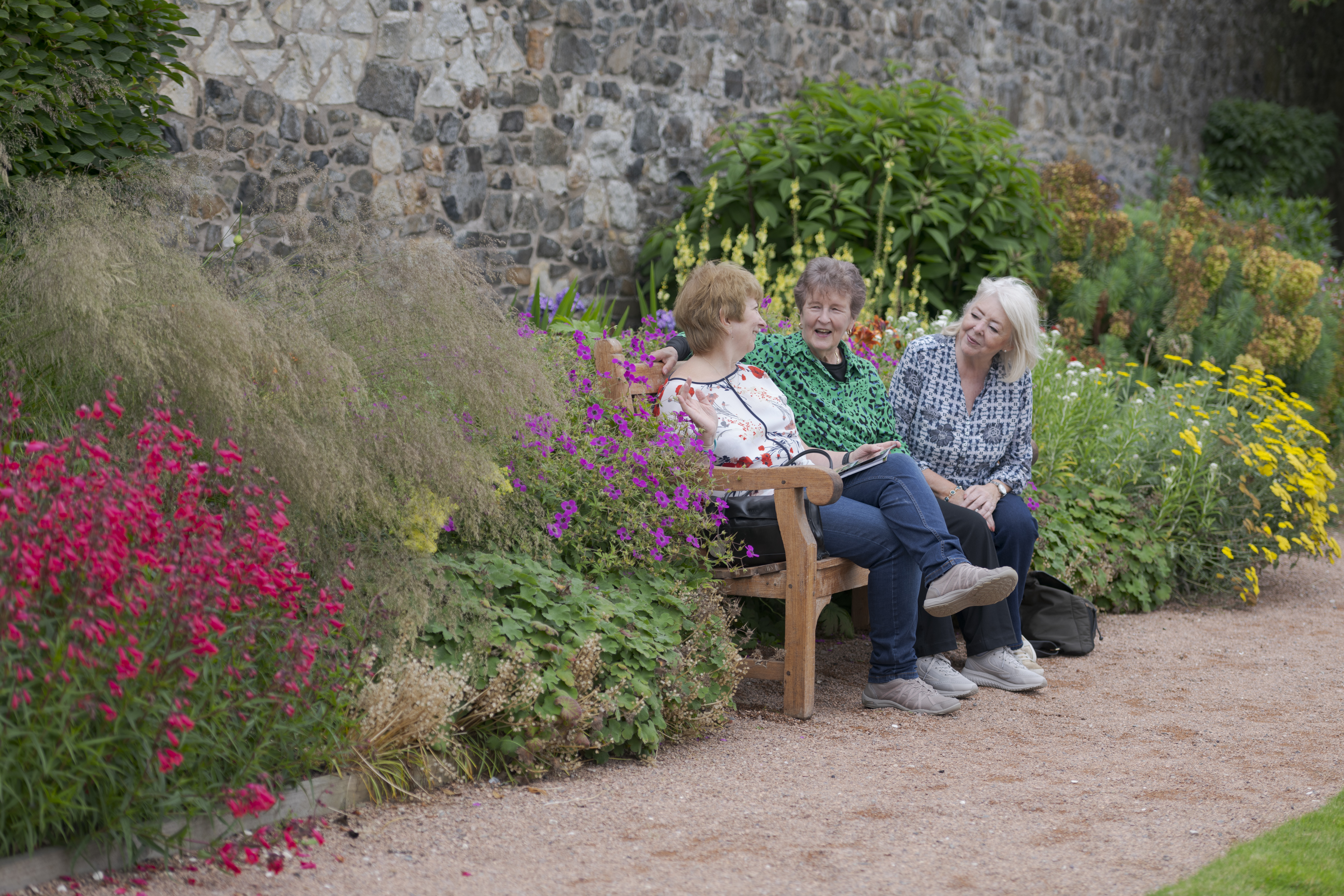 Three visitors sit on a wooden bench in a colourful garden, chatting among flowering plants beside a stone wall.