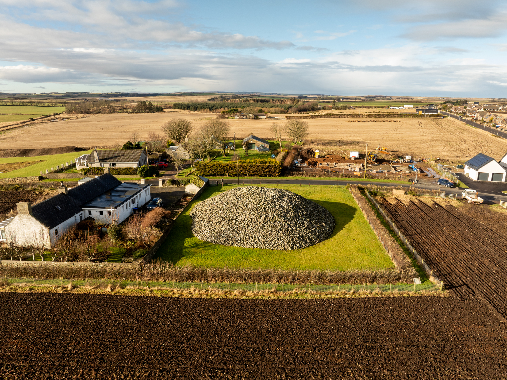 Aerial photograph showing Memsie Cairn in a green enclosure, surrounded by farmland, houses, and construction activity in the distance