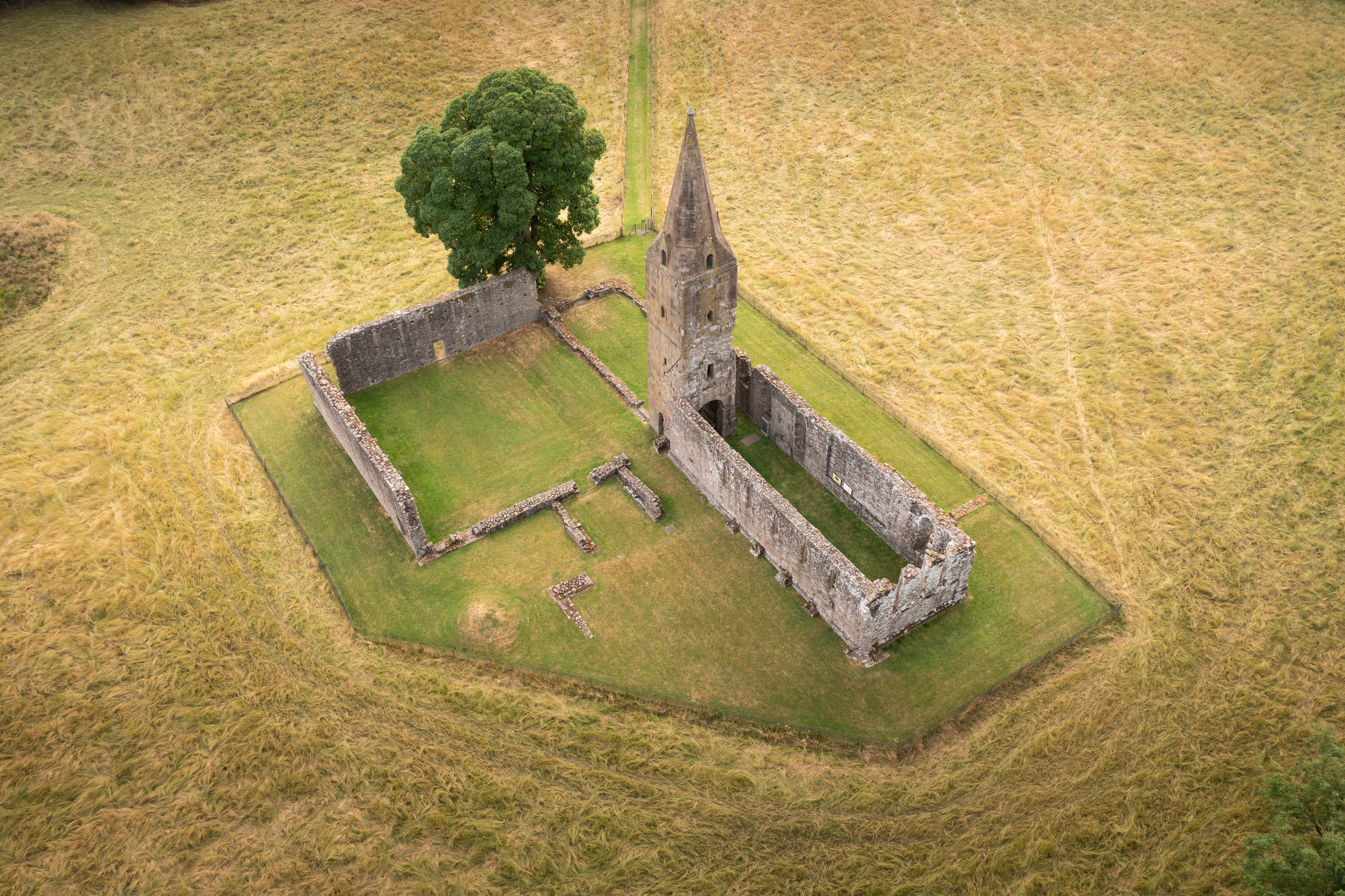 Parched grass creates a yellow sea with a green island in the middle of it, neatly shaped to accommodate the ruins of a priory with a tower still in tact