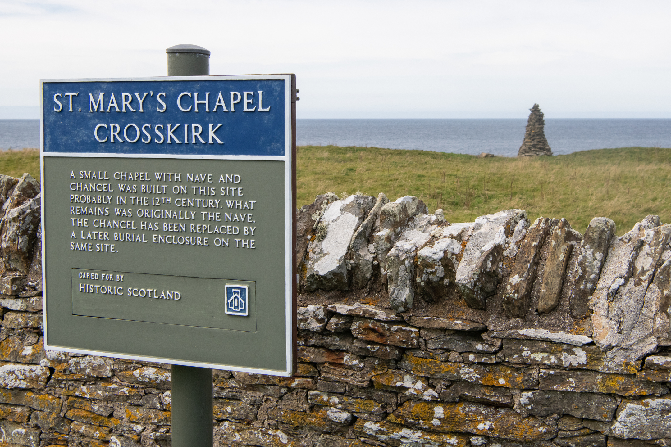 A close view of an informational sign mounted on a stone wall. The sign reads “St. Mary’s Chapel Crosskirk,” followed by a historical description of the site. Behind the wall stretches a grassy field leading toward rocky coastal cliffs and the open sea. A tall cairn is visible in the distance near the shoreline. The sky is lightly overcast