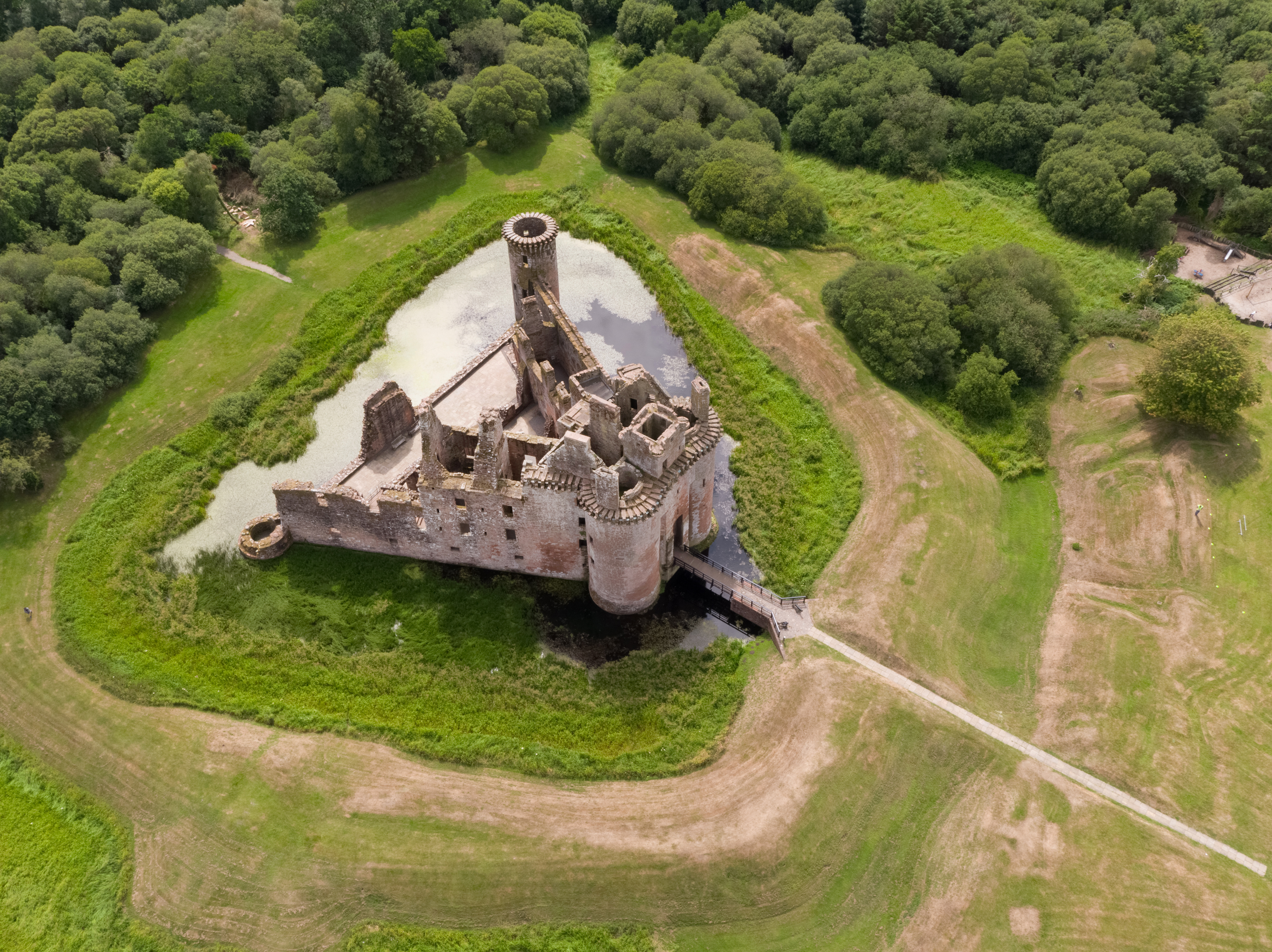 Aerial view of a ruined stone castle set out in a triangular shape and surrounded by a water‑filled moat. The castle’s walls and towers are roofless and partially collapsed, with a tall circular tower rising at one corner. A narrow bridge crosses the moat to the entrance.
