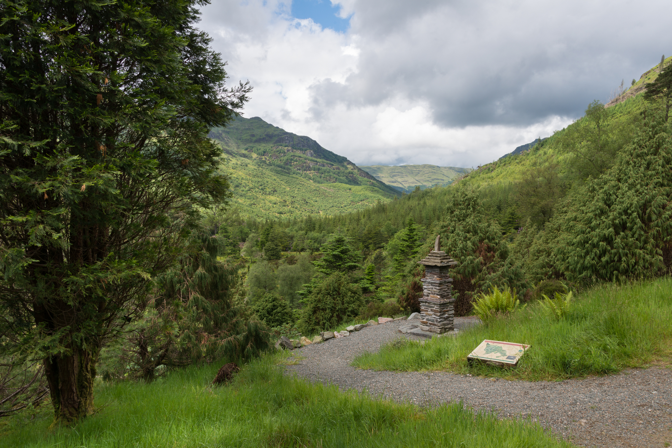 Scenic green mountain valley with dense woodland, rolling hills, and a small stone monument beside a gravel path under a cloudy sky.