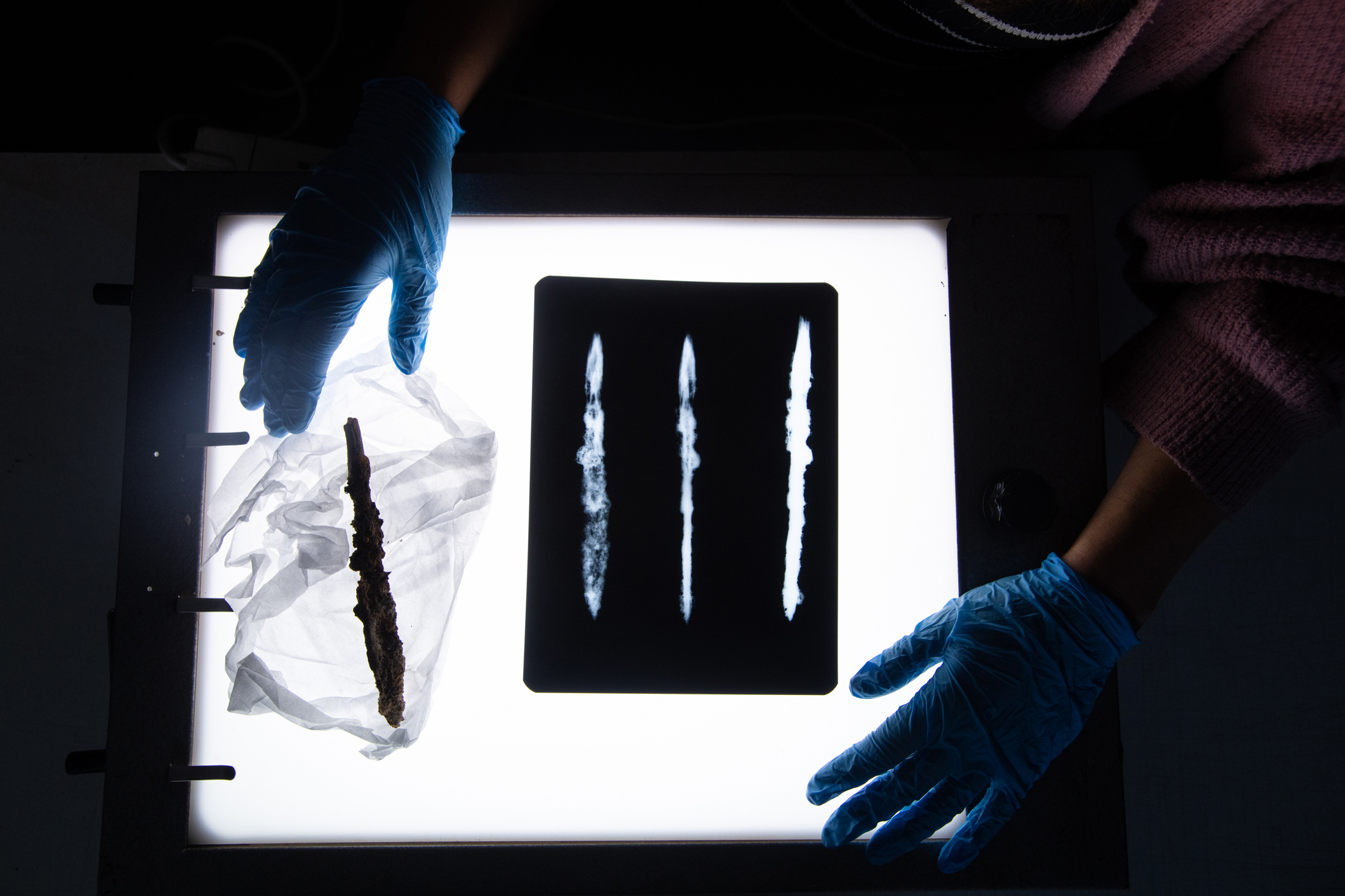 An x-ray of a Viking arrowhead on a lightbox. A person with gloved hands stands over the box.