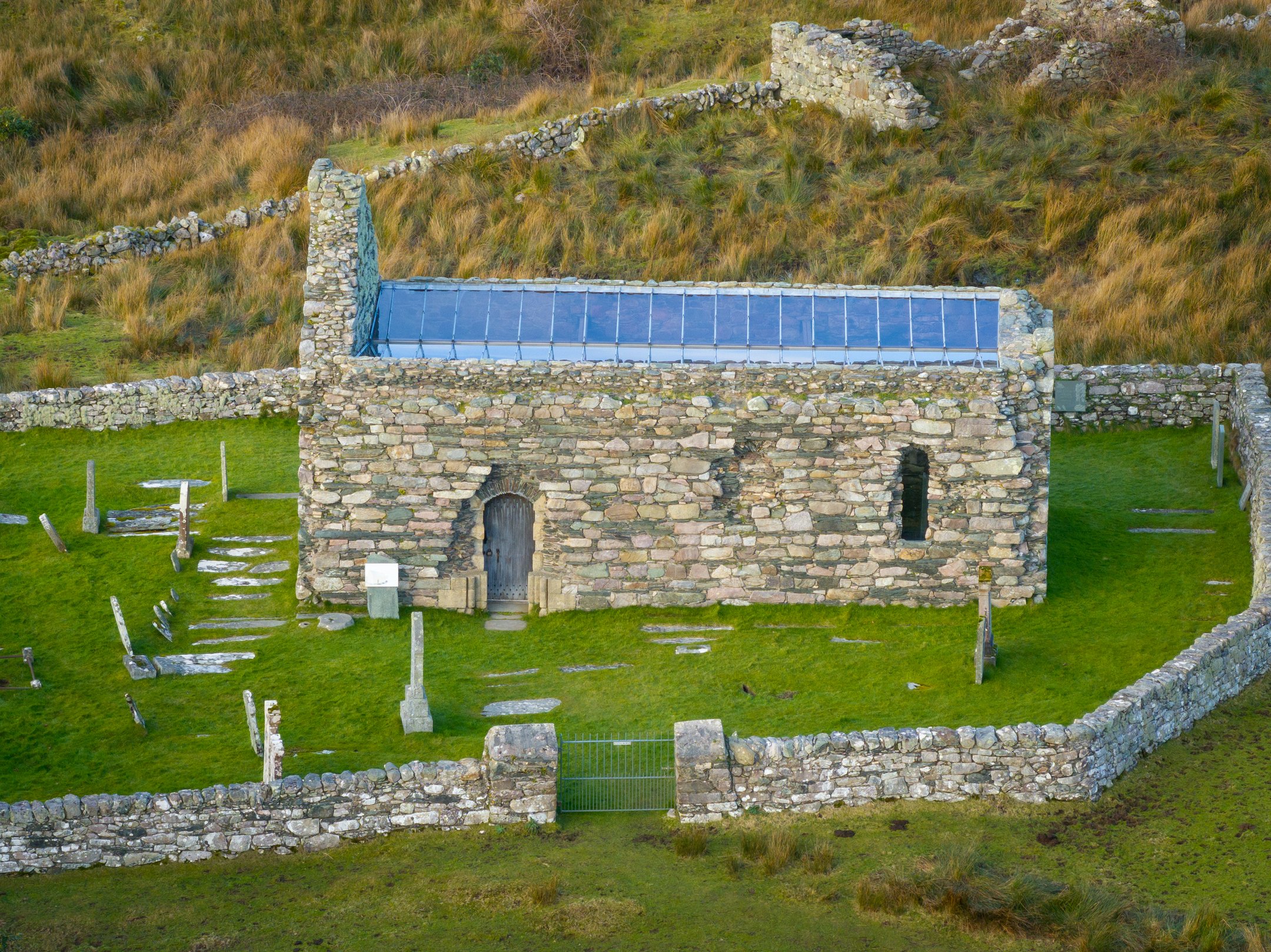 An old rectangular stone chapel with a wooden door and a modern transparent roof, surrounded by a walled graveyard on grassy ground.