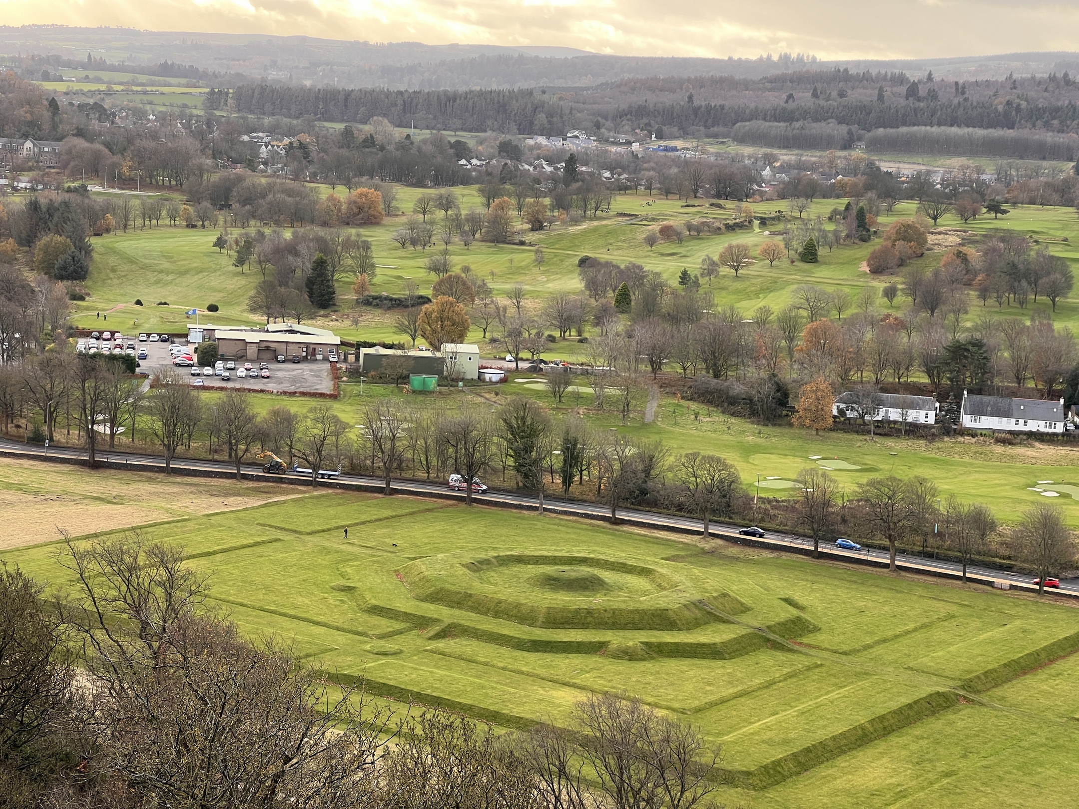 Aerial-style view of the King’s Knot’s distinctive terraced, octagonal mound set within a large rectangular field. The surrounding area includes scattered buildings, trees, and sweeping green hills beneath soft golden light.