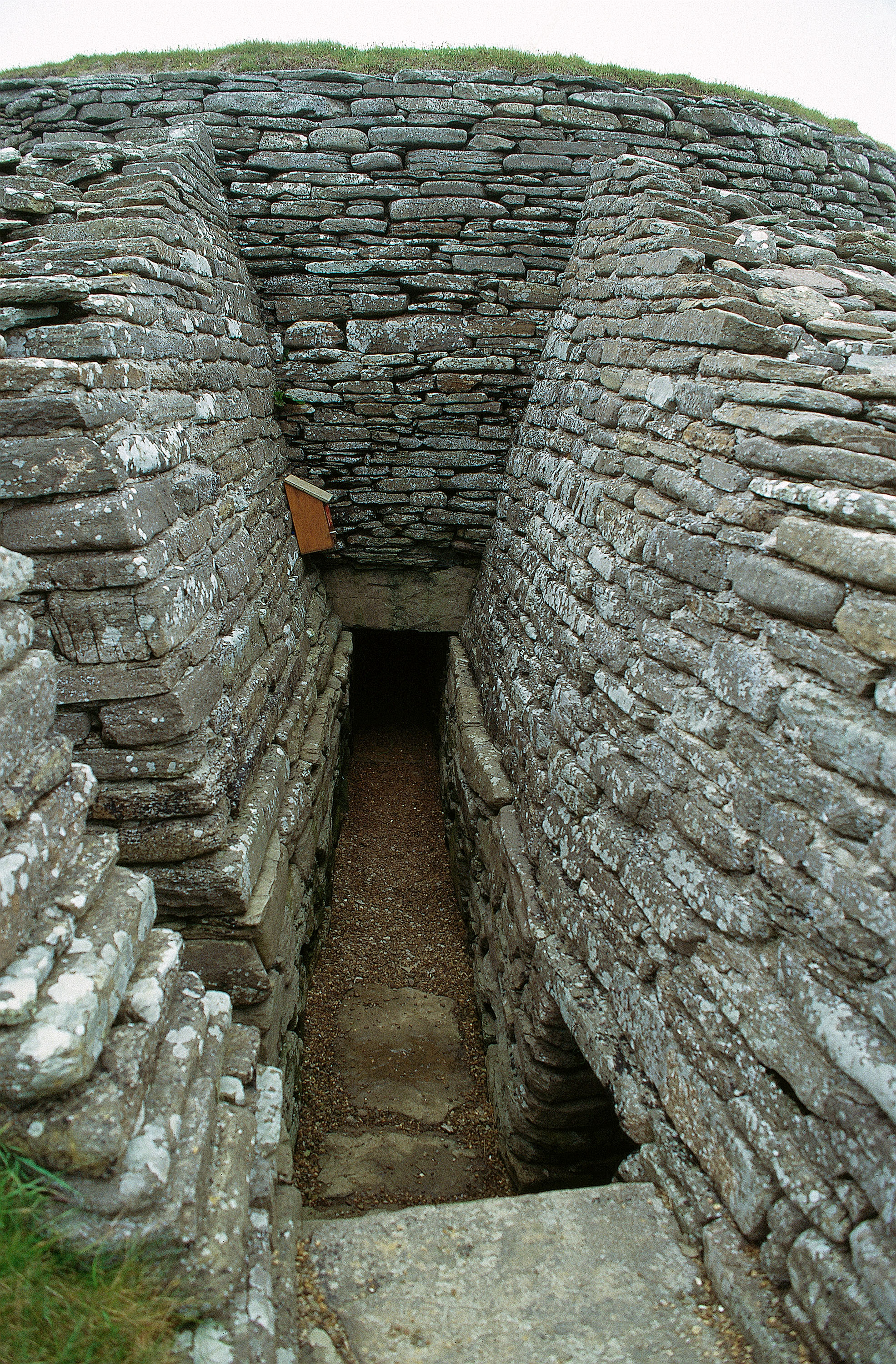 Entrance passage of Quoyness Chambered Cairn, showing a long, narrow drystone corridor built from tightly stacked stone slabs. The passage slopes gently downward toward a dark doorway covered by a large horizontal capstone. The high stone walls curve inward slightly as they lead into the mound, with patches of lichen visible on the stones.