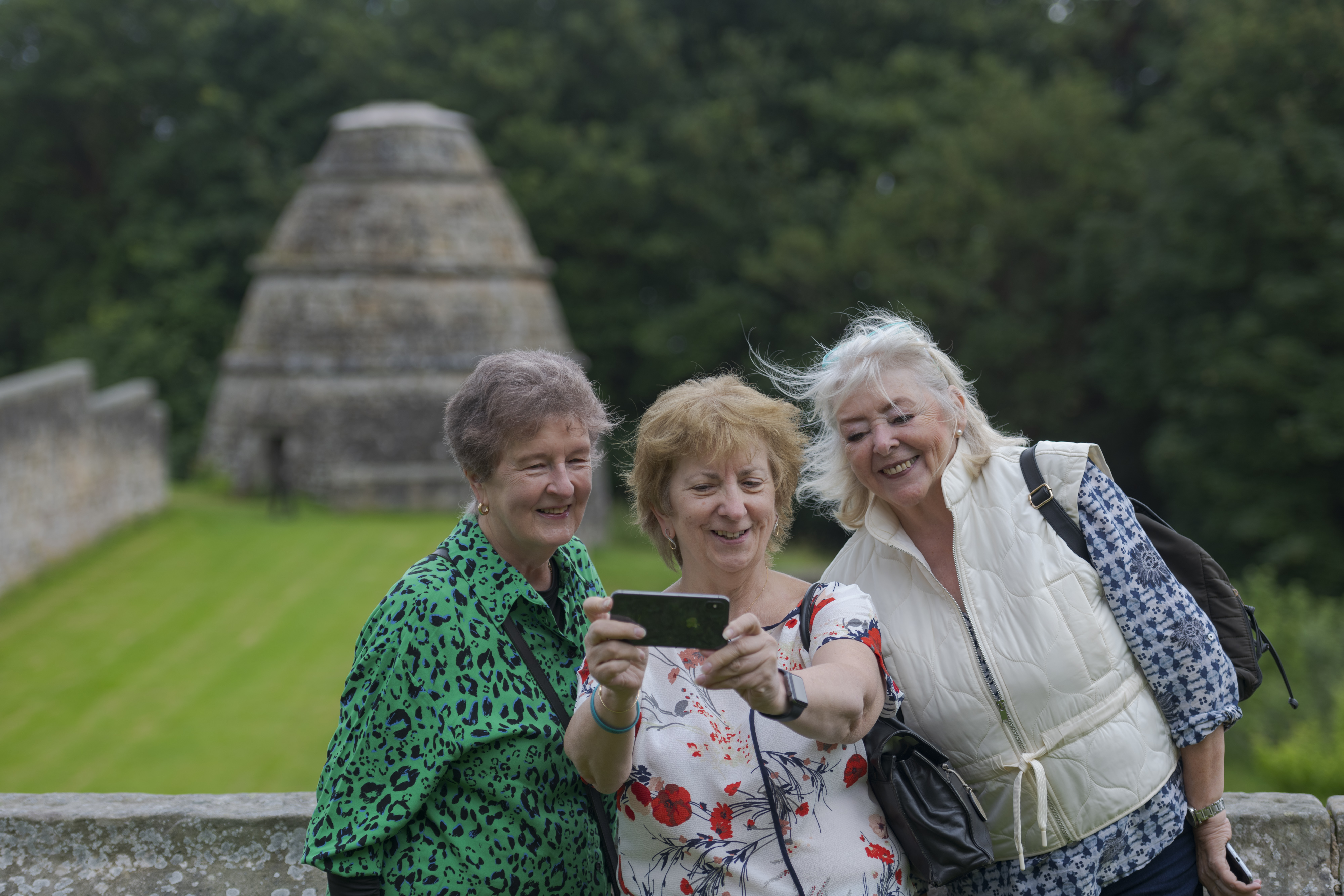 Three visitors stand close together, taking a selfie, with a historic stone dovecot structure in the background.