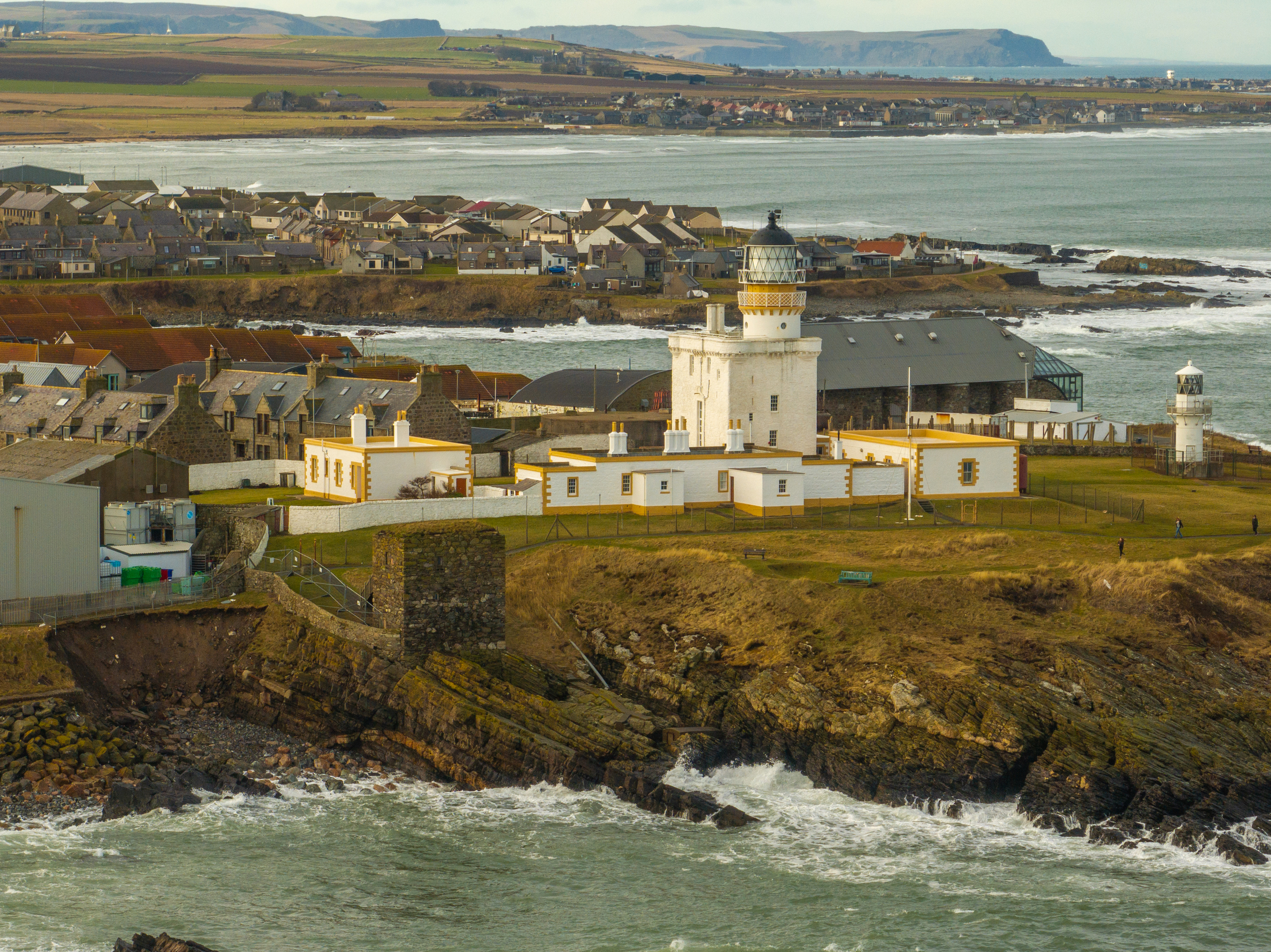 An aerial view of a historic lighthouse complex. A square, castle-like building is topped with a large circular light. It is surrounded by various ancillary buildings, All the walls are whitewashed with a pleasant yellow paint trim.