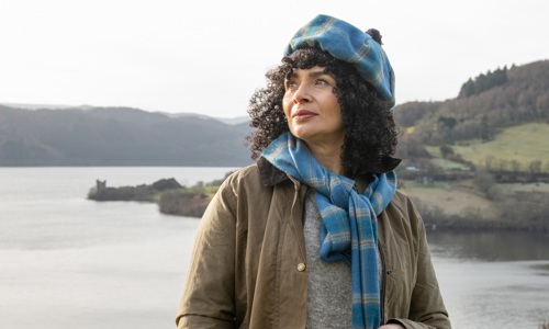 A woman with dark brown curly hair wears a hat and scarf with a blue and grey tartan pattern. The model is wearing a brown jacket and is looking off to her right-hand side in the distance. In the background there is a castle, trees and grass and a large loch.