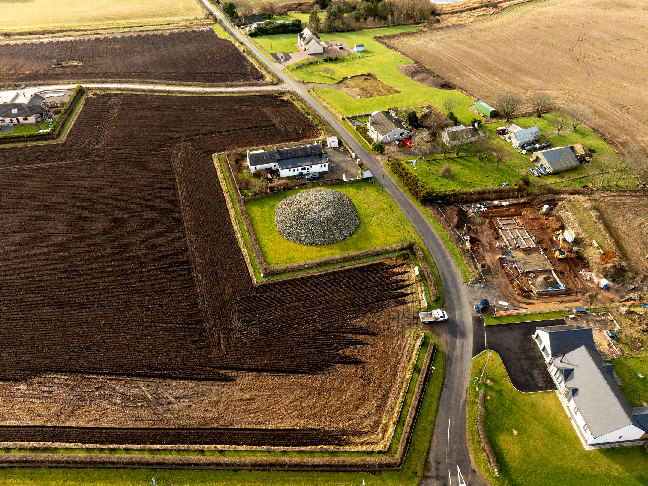 Aerial view of Memsie Cairn, a large circular stone mound surrounded by green grass, located near farm fields and houses in a rural landscape.