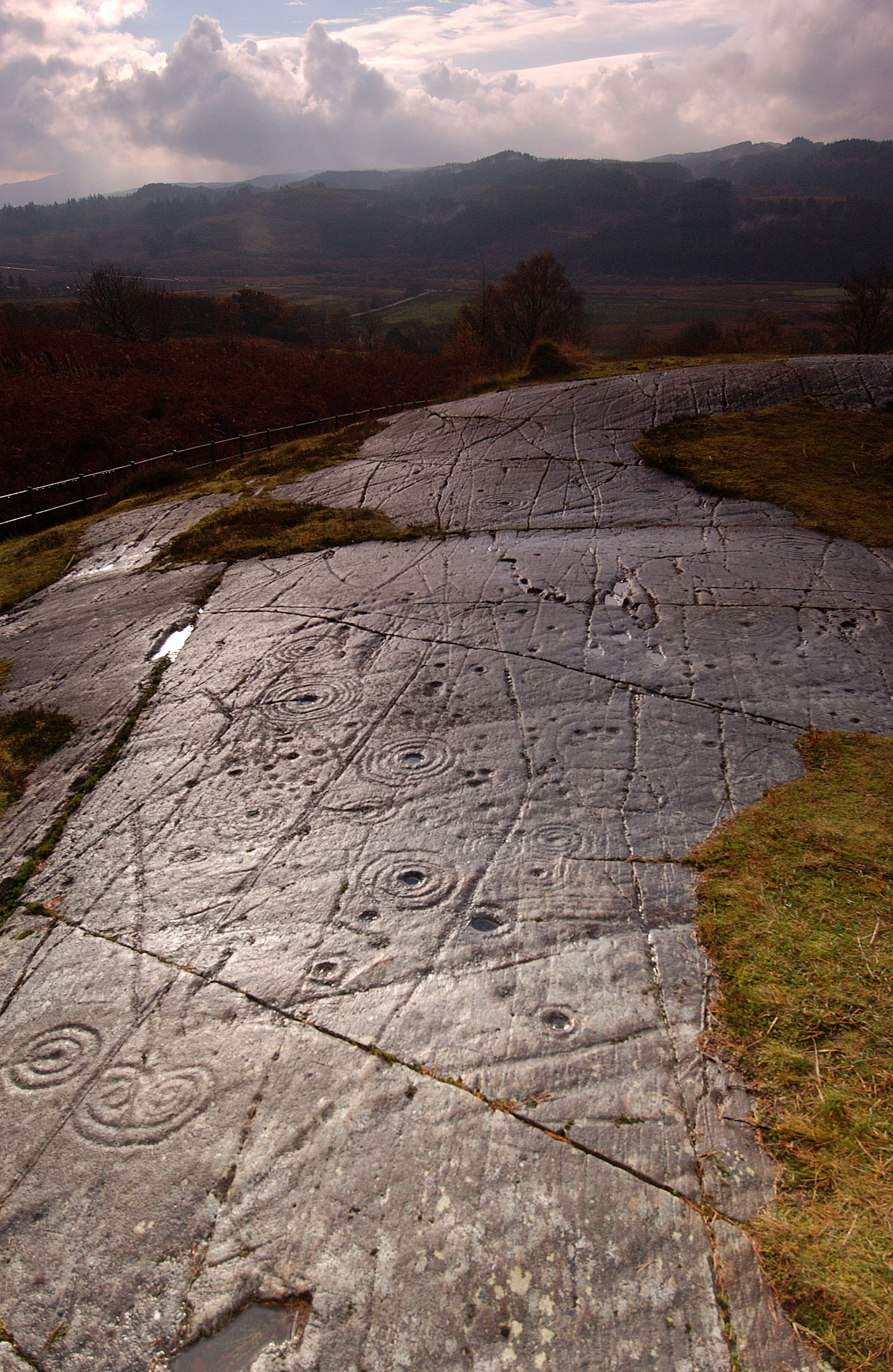 Flat rock with concentric circle carvings and grooves, surrounded by grass and hills under cloudy sky.