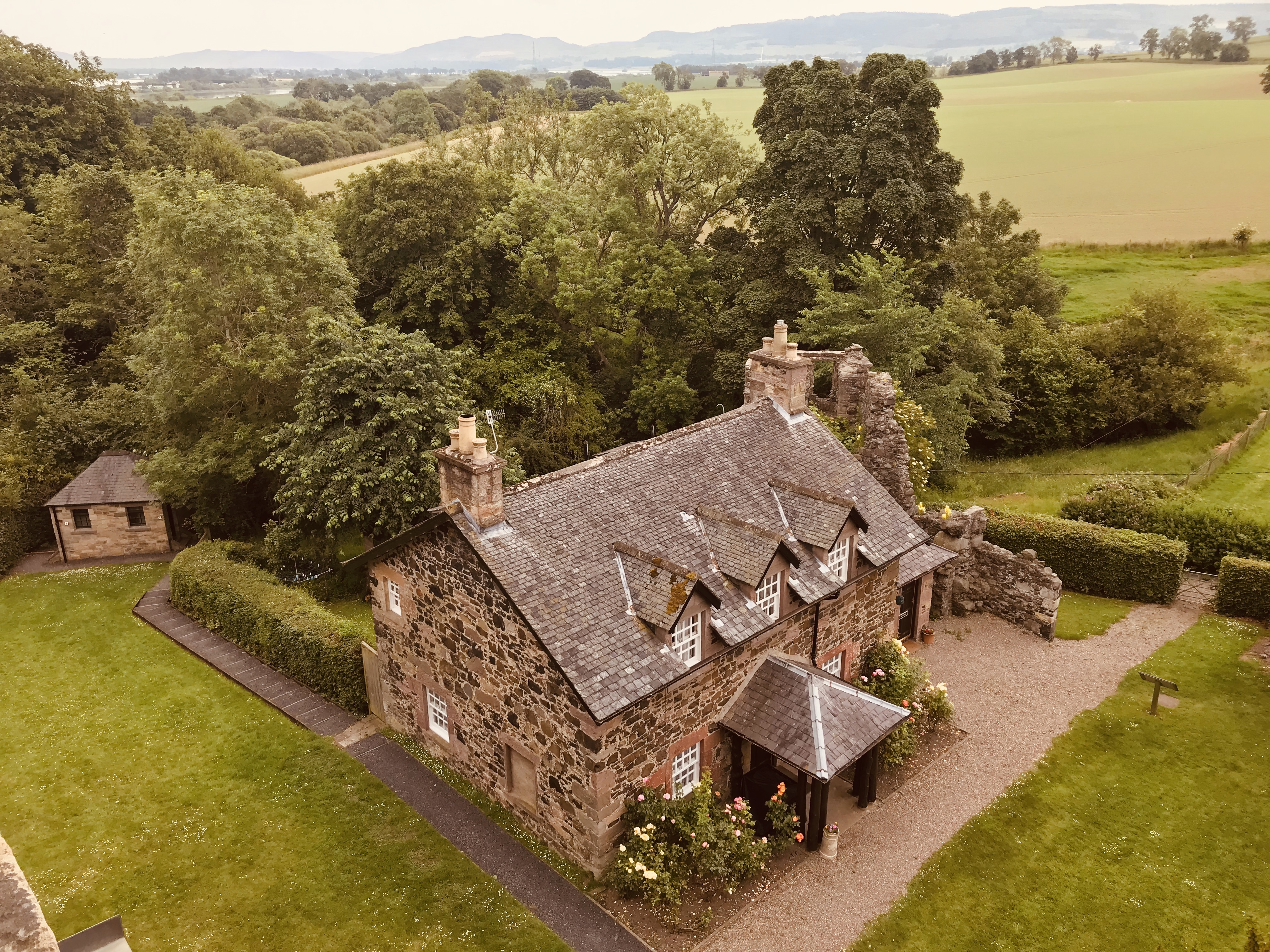 Aerial view of a stone-built cottage with a slate roof and dormer windows, set within a green garden beside Elcho Castle. Trees and open countryside stretch into the distance under a hazy sky.