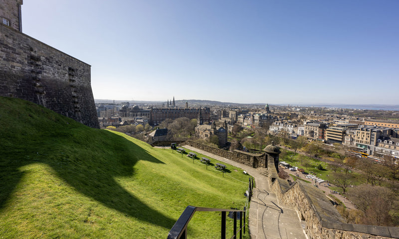 A brick wall at the top of a hill of grass with steps to walk up/down and a view over a city with bright blue skies