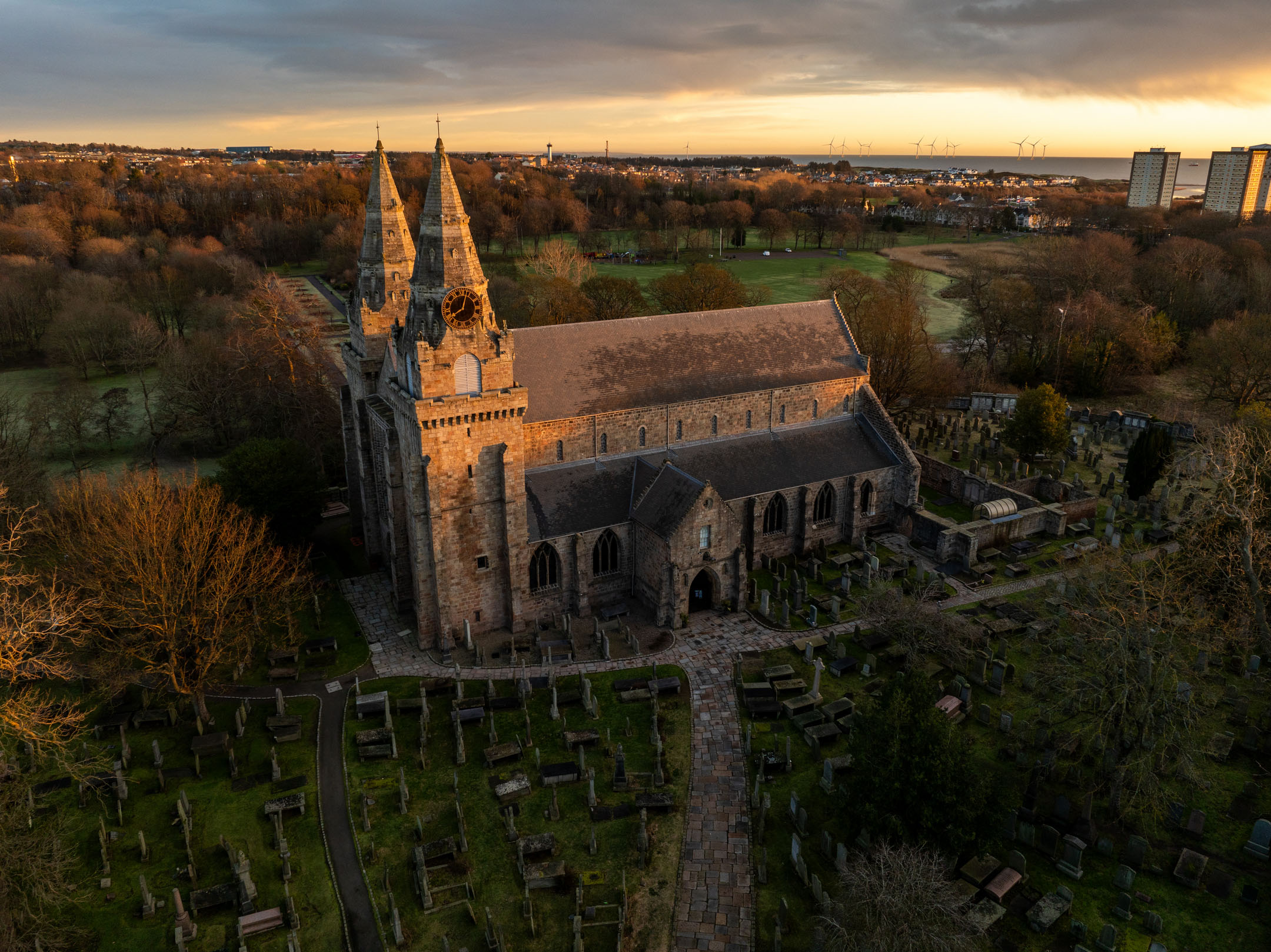 Aerial view of St Machar's Cathedral at sunset with twin spires and surrounding graveyard.