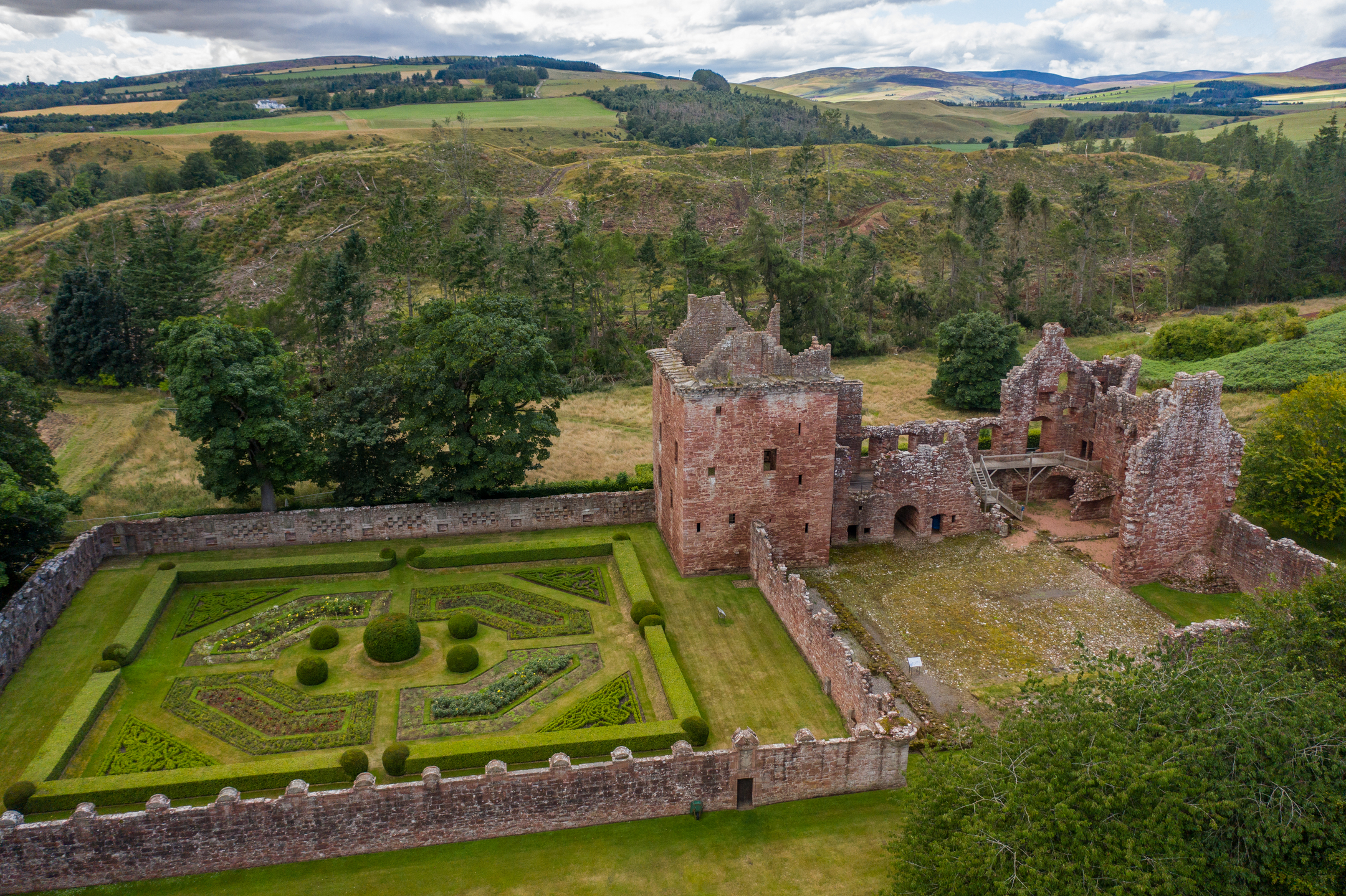An aerial photo of a ruined castle surrounded by green trees. Beside the castle is an ornate, symmetrical walled garden. 