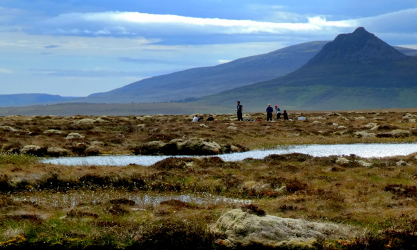 Dubh Lochans below Maiden Pap. A boggy, marsh landscape with a mountain range in the background. There is a group of people in the mid-distance. 