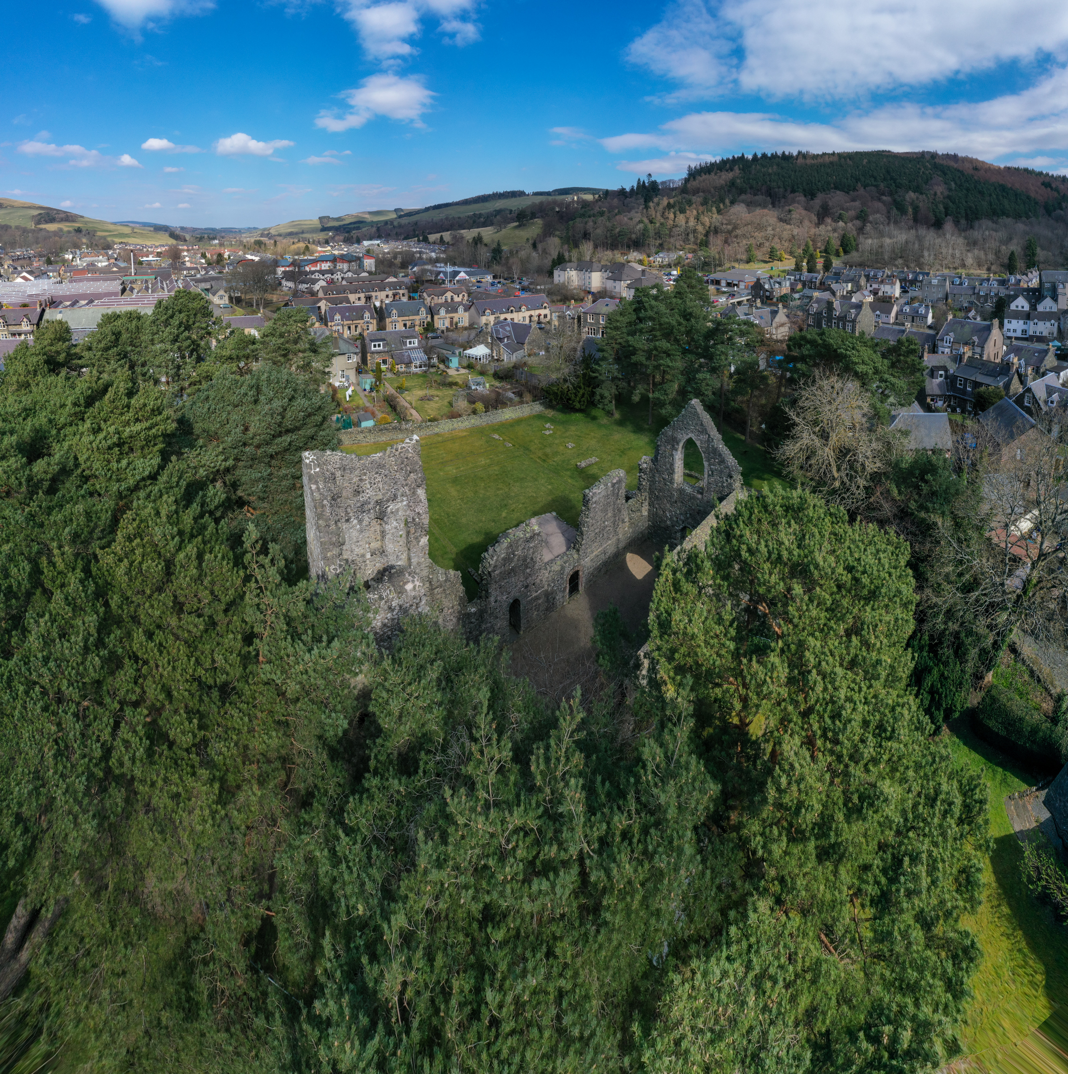 Elevated view of Cross Kirk ruins partially hidden among tall green trees. The stone walls rise from a grassy area, with the town of Peebles and rolling hills visible in the distance.