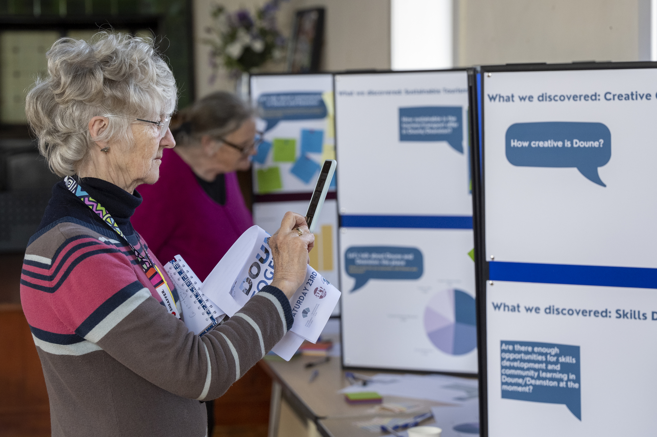 A lady at a community event uses her phone to take a photo of the information displayed on a notice board.
